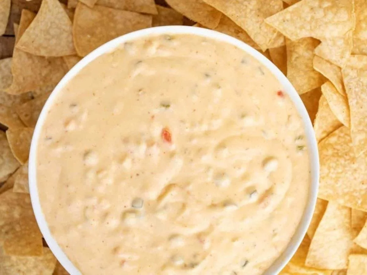 A white bowl filled with creamy white queso dip containing bits of peppers, surrounded by a bed of crunchy yellow tortilla chips.