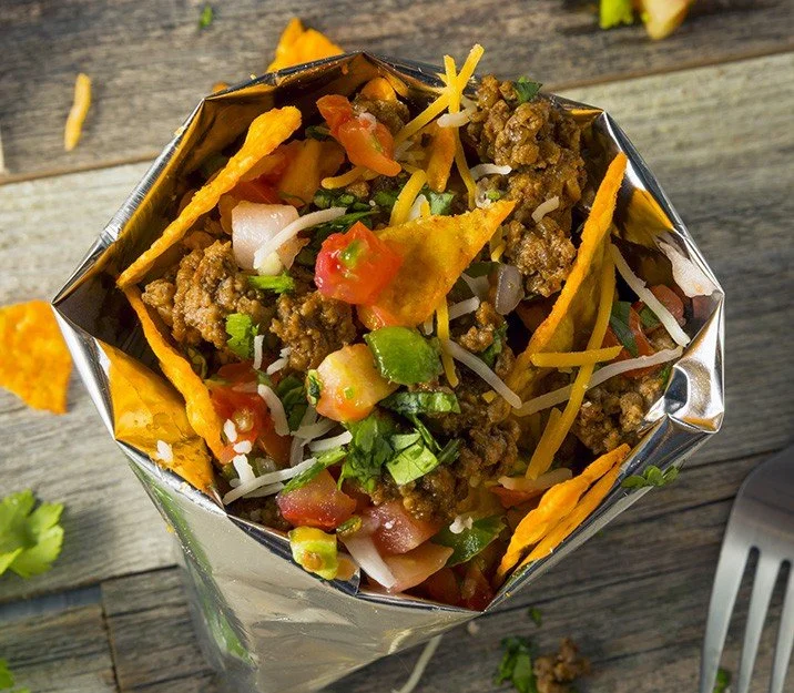 A walking taco served in a silver foil bag, filled with seasoned ground beef, shredded cheddar cheese, diced tomatoes, onions, and fresh cilantro over crunchy corn chips.