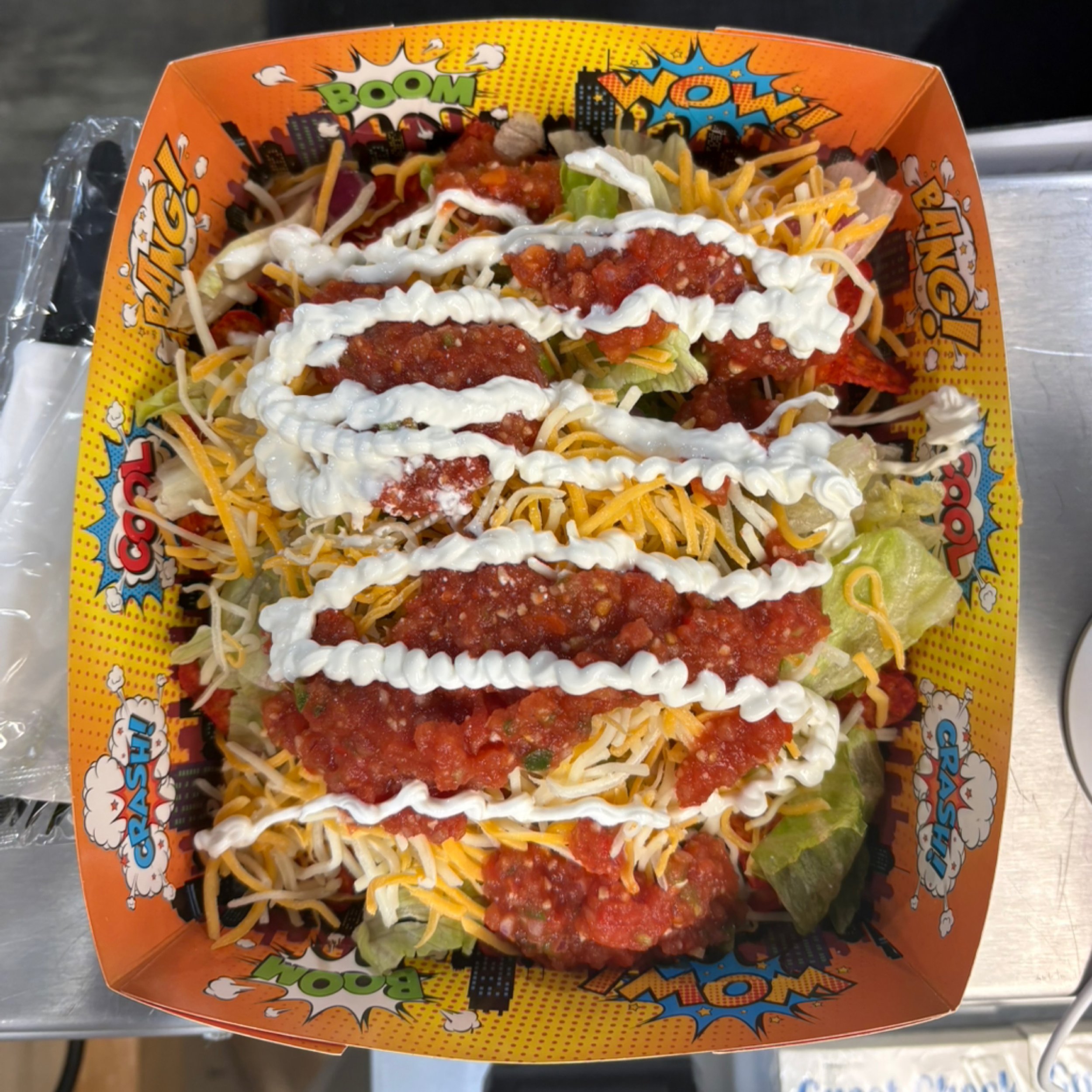 Overhead view of a "Build Your Own" nacho bowl. The dish is layered with fresh lettuce, a generous layer of shredded cheese, rows of red salsa, and zig-zags of white sour cream.