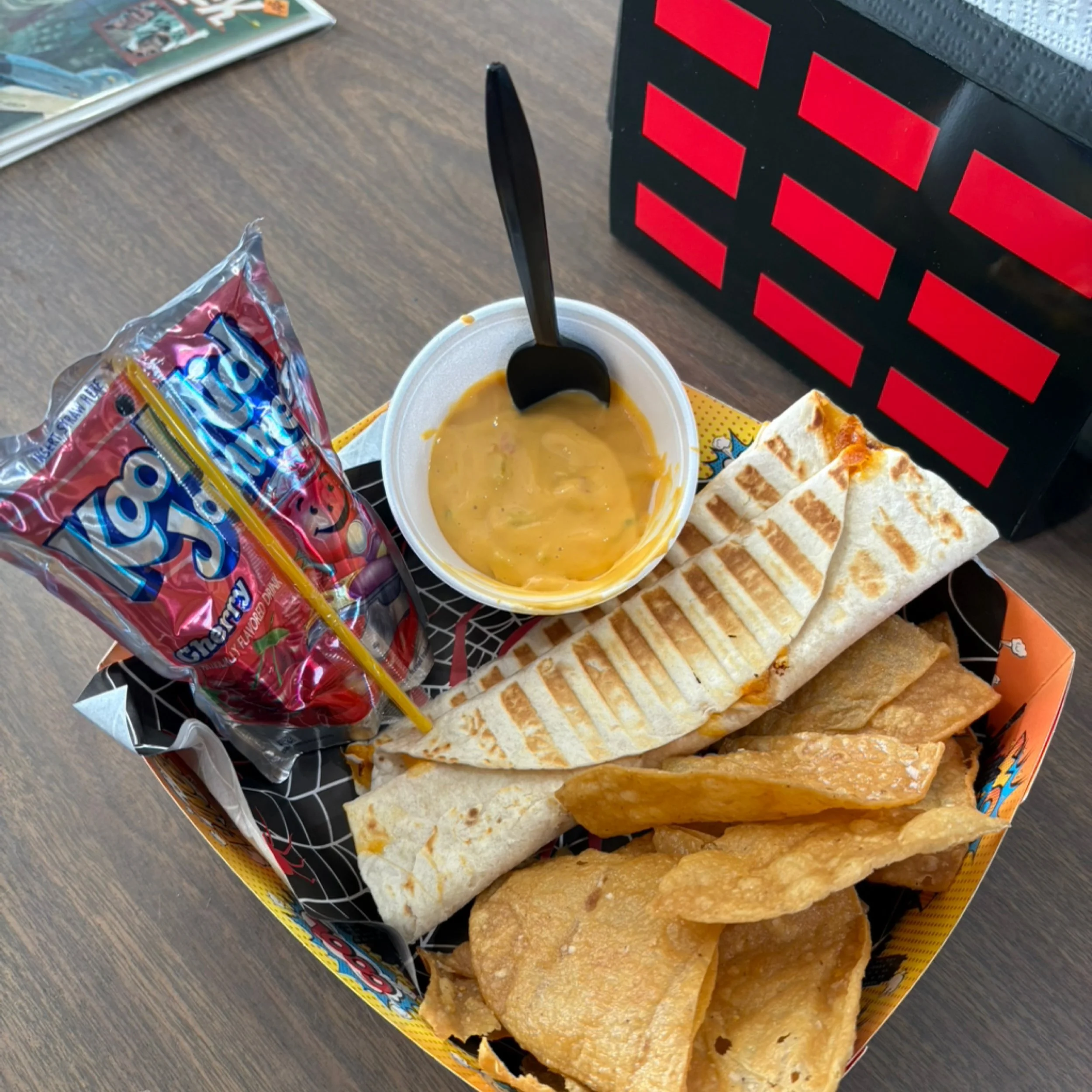 A burrito meal featuring a toasted, grill-marked burrito, a side of tortilla chips, a small cup of nacho cheese sauce, and a cherry Kool-Aid Jammers pouch.