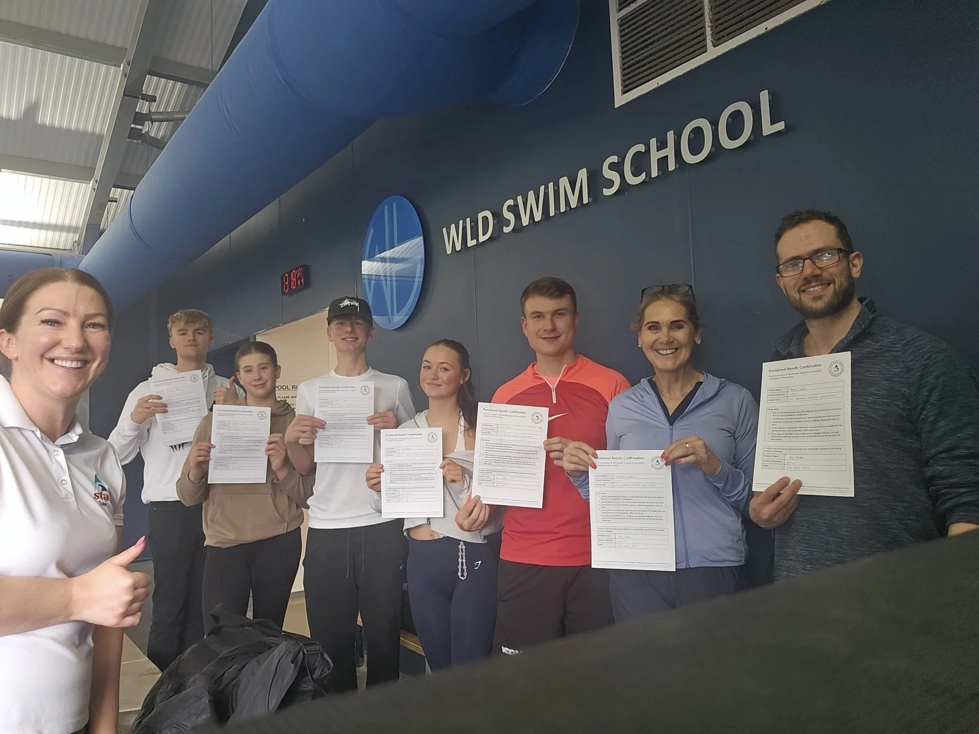 Group of people holding certificates at WLD Swim School, indoor pool area, smiling, some wearing casual athletic clothing, background features a blue wall with a logo and the name of the swim school.