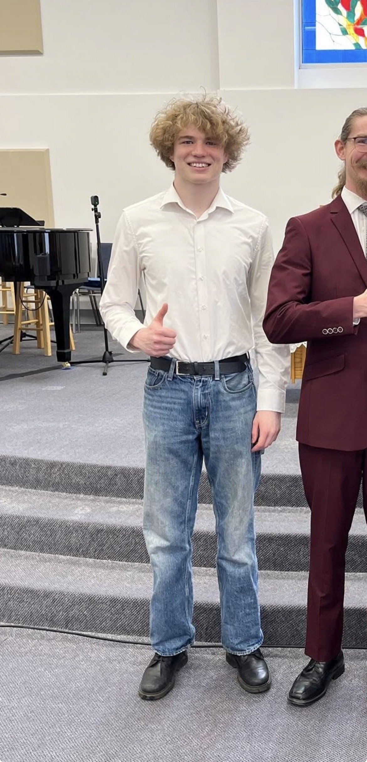 A young man with curly blond hair, wearing a white button-down shirt and jeans, smiling and giving a thumbs-up. He is standing on a carpeted staircase inside a church or community hall, with a piano and chairs in the background.