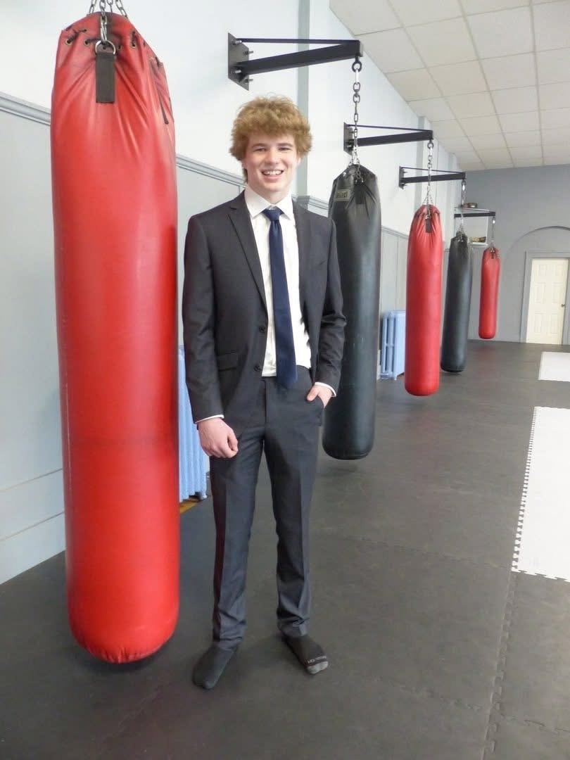 A young man in a black suit, white shirt, and navy tie standing inside a boxing gym, smiling, with several hanging punching bags behind him.