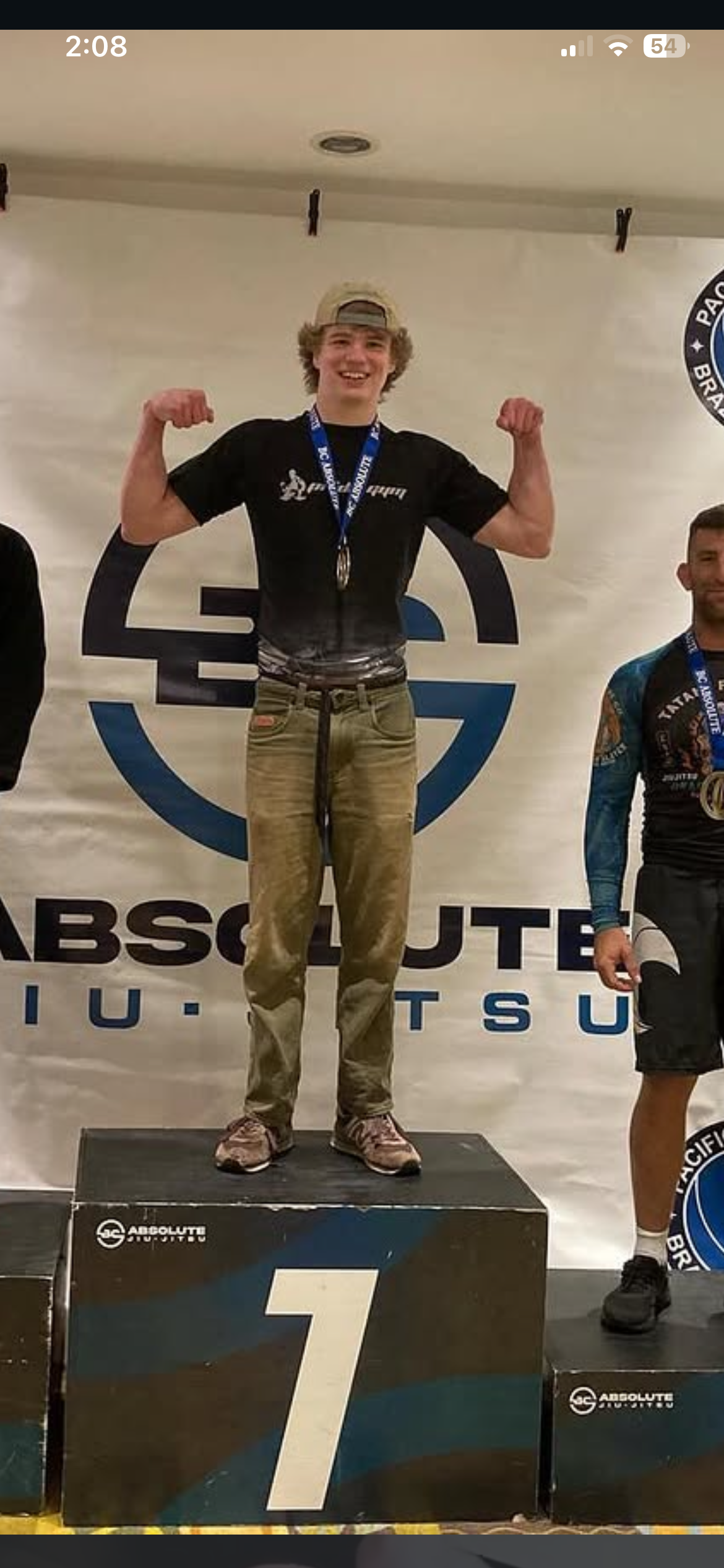 Young man standing on a first-place winner's podium, flexing his arms in a victory pose, wearing a black T-shirt, beige pants, a backwards cap, and a medal around his neck, at a martial arts competition.
