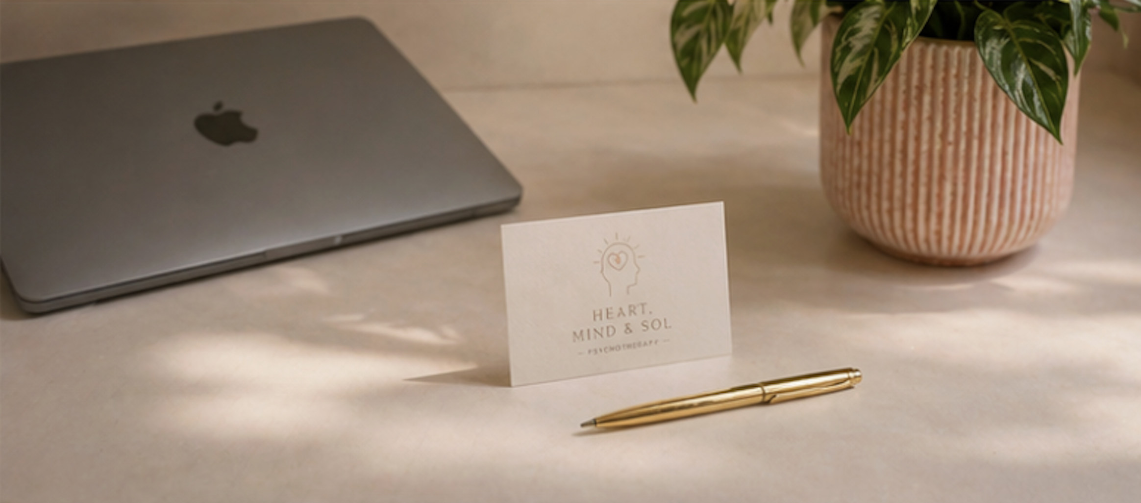 A workspace with a closed silver Apple laptop, a pink textured planter with green and white variegated leaves, a white card with a human profile and heart logo, and a gold pen, all on a light-colored surface.