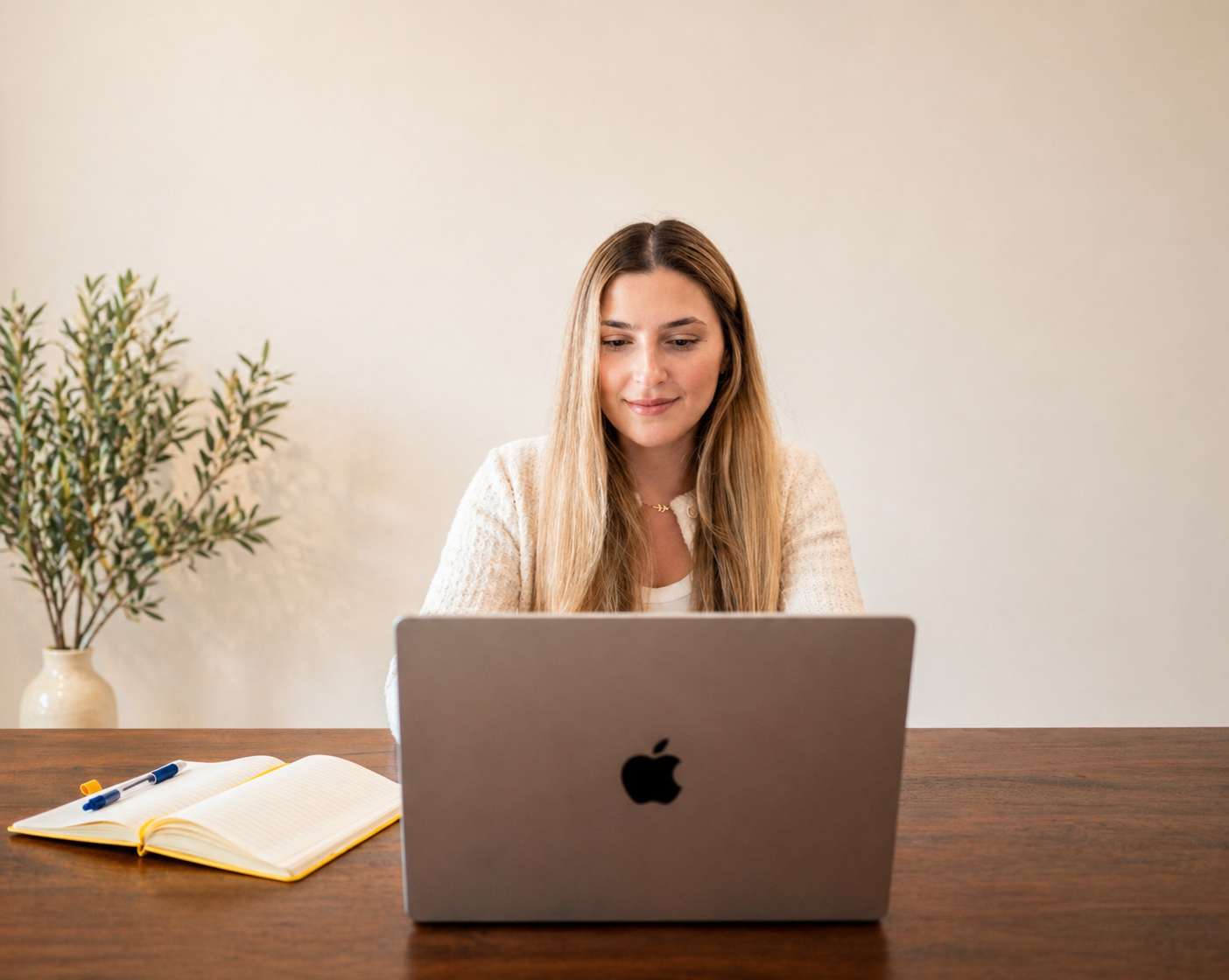 A woman with long blonde hair working on a laptop at a wooden desk, with a potted plant in a vase and an open notebook with pens nearby.