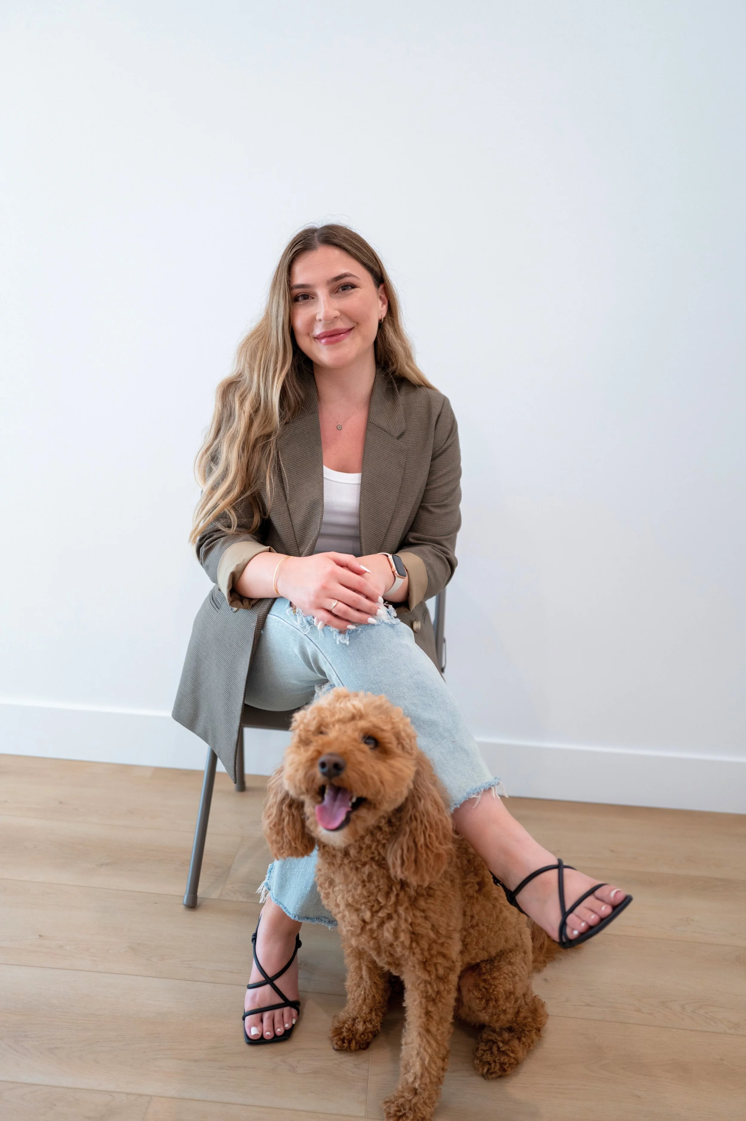 A woman with long wavy hair and a smile, sitting on a gray chair on a wooden floor, with a brown poodle in front of her. She is wearing a brown blazer, ripped light blue jeans, black strappy sandals, and a smartwatch. The background is a plain white wall.