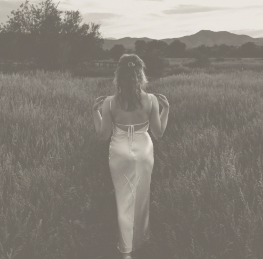 A woman in a white dress walking away through a grassy field at sunset or sunrise.