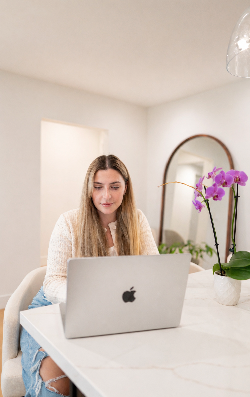 A young woman with long blonde hair, wearing a cream-colored sweater, sitting at a white table, looking at a silver Apple laptop. A purple orchid in a white pot is on the table, and a round mirror hangs on the white wall behind her.