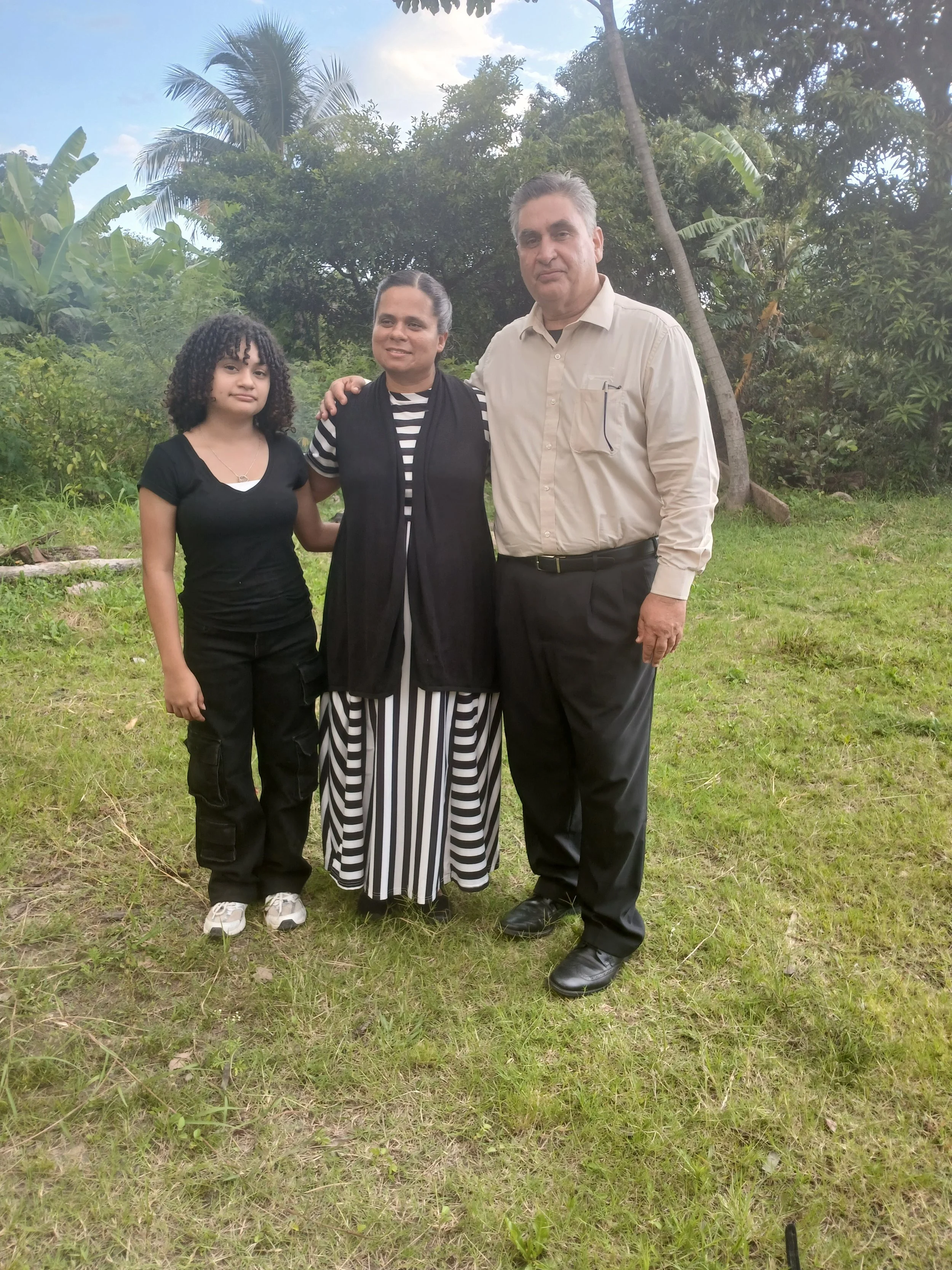 A group of three people standing outdoors on green grass, with a backdrop of trees and a partly cloudy sky. The group includes a young girl, a middle-aged woman, and a middle-aged man, posing together for a photo.
