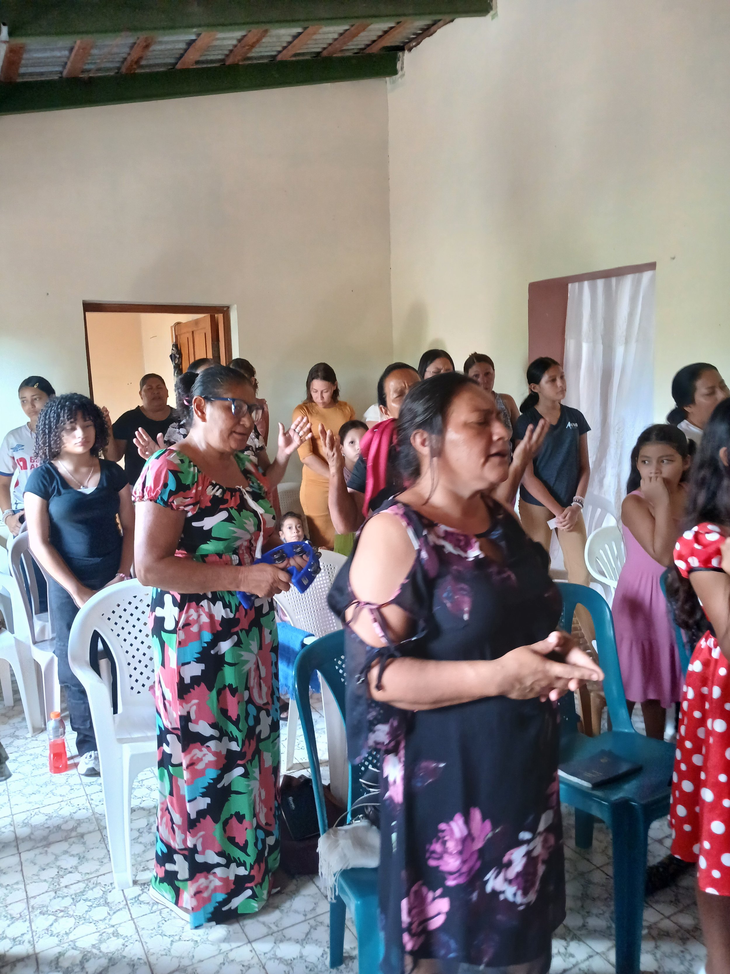 A group of people, mostly women and children, gathered indoors, some with eyes closed and hands raised, possibly praying or participating in a religious or community event.