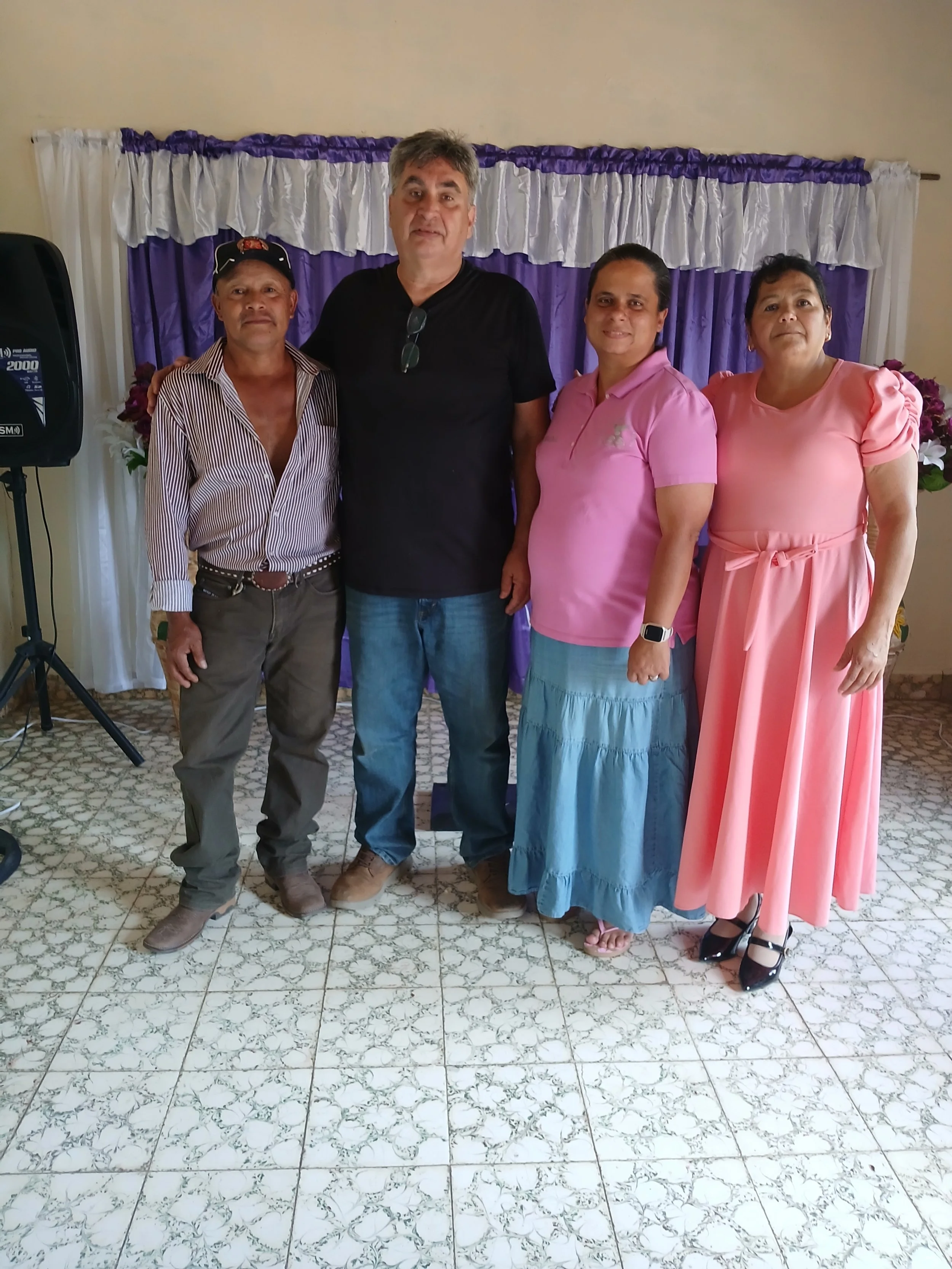Four people standing together in front of a purple and white curtain, posing for a photo indoors. Two women on the right dressed in pink, one man in a striped shirt and cap on the left, and a tall man in a black shirt in the middle.