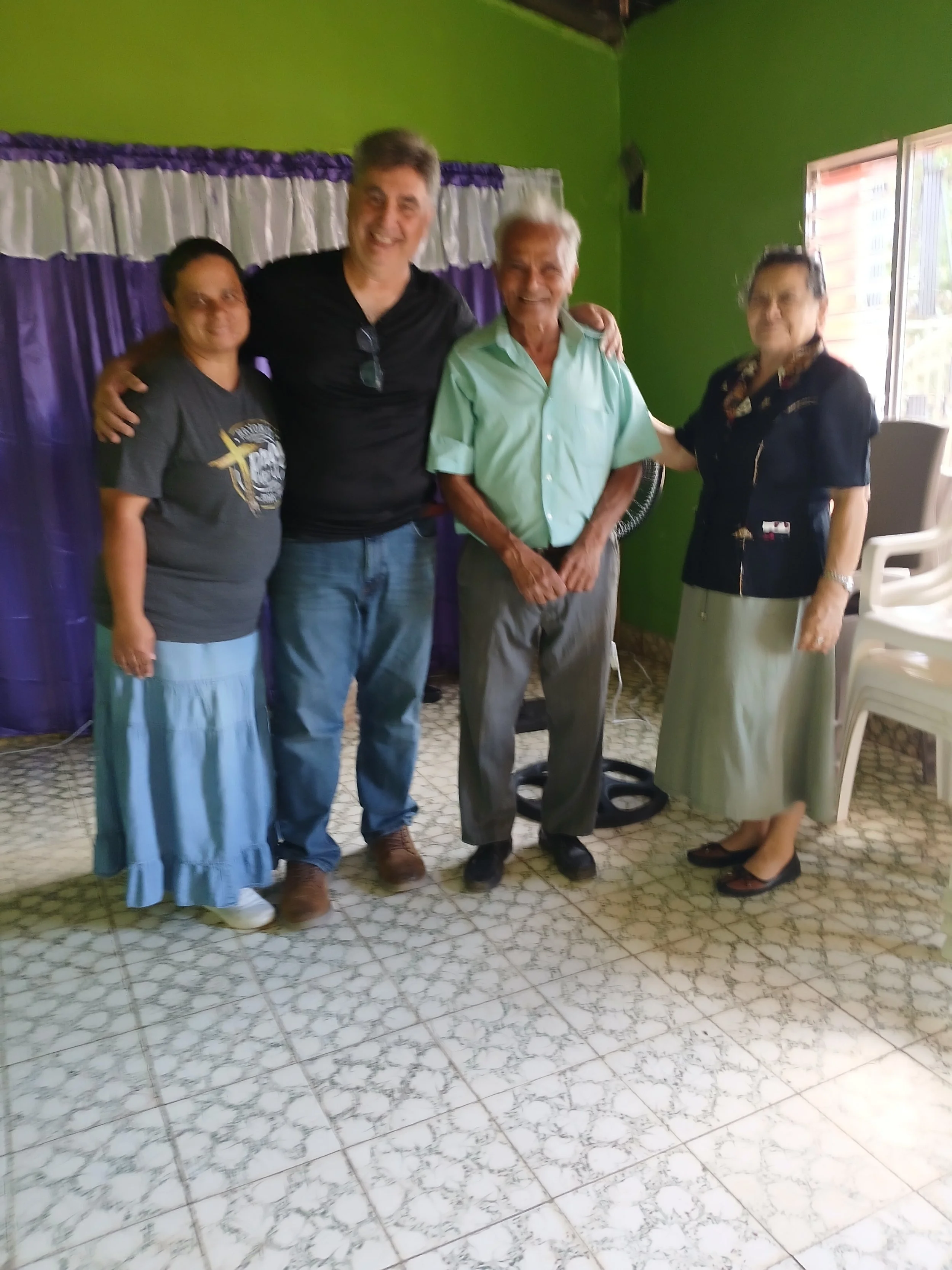 Four people standing together indoors, smiling, with purple curtains and green walls behind them, and a tiled floor.