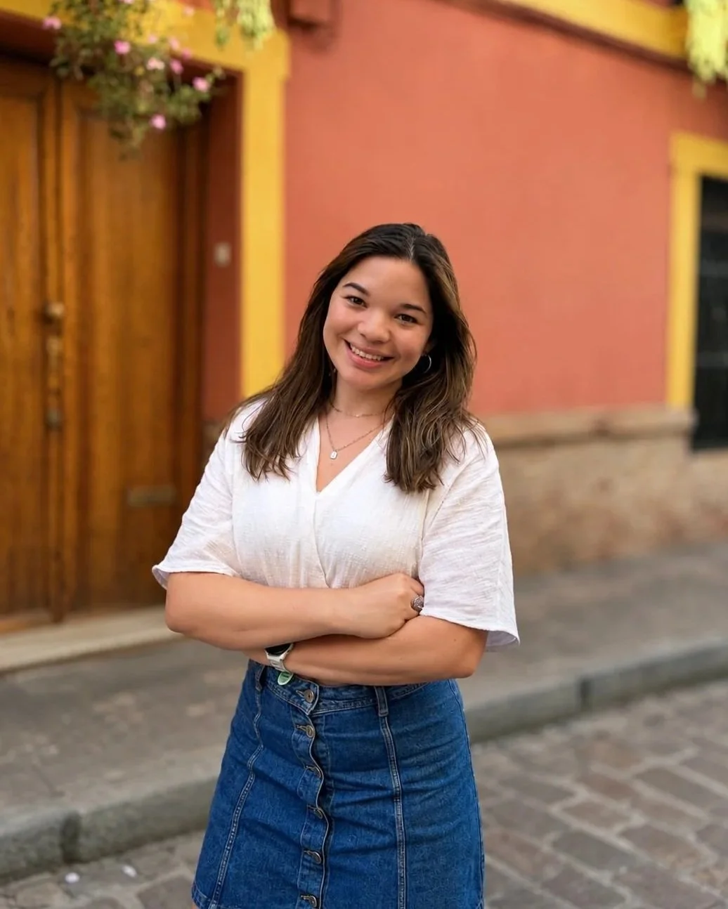 A young woman with shoulder-length brown hair, wearing a white V-neck blouse and a denim skirt, smiling with crossed arms in front of colorful buildings with pink and yellow walls and wooden door.