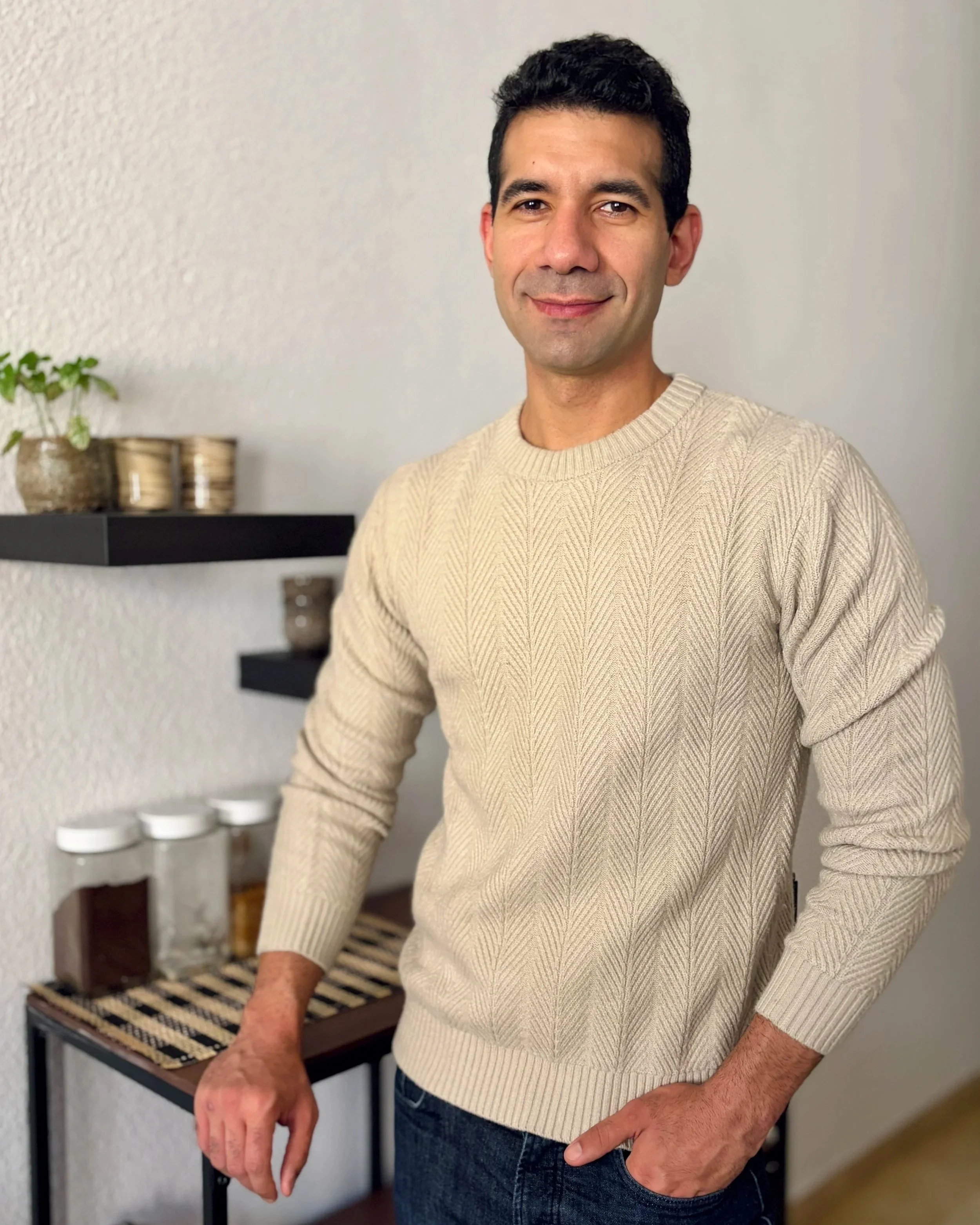 Midsection of a man wearing a beige cable-knit sweater, standing indoors near a black shelf with potted plants and jars in the background.