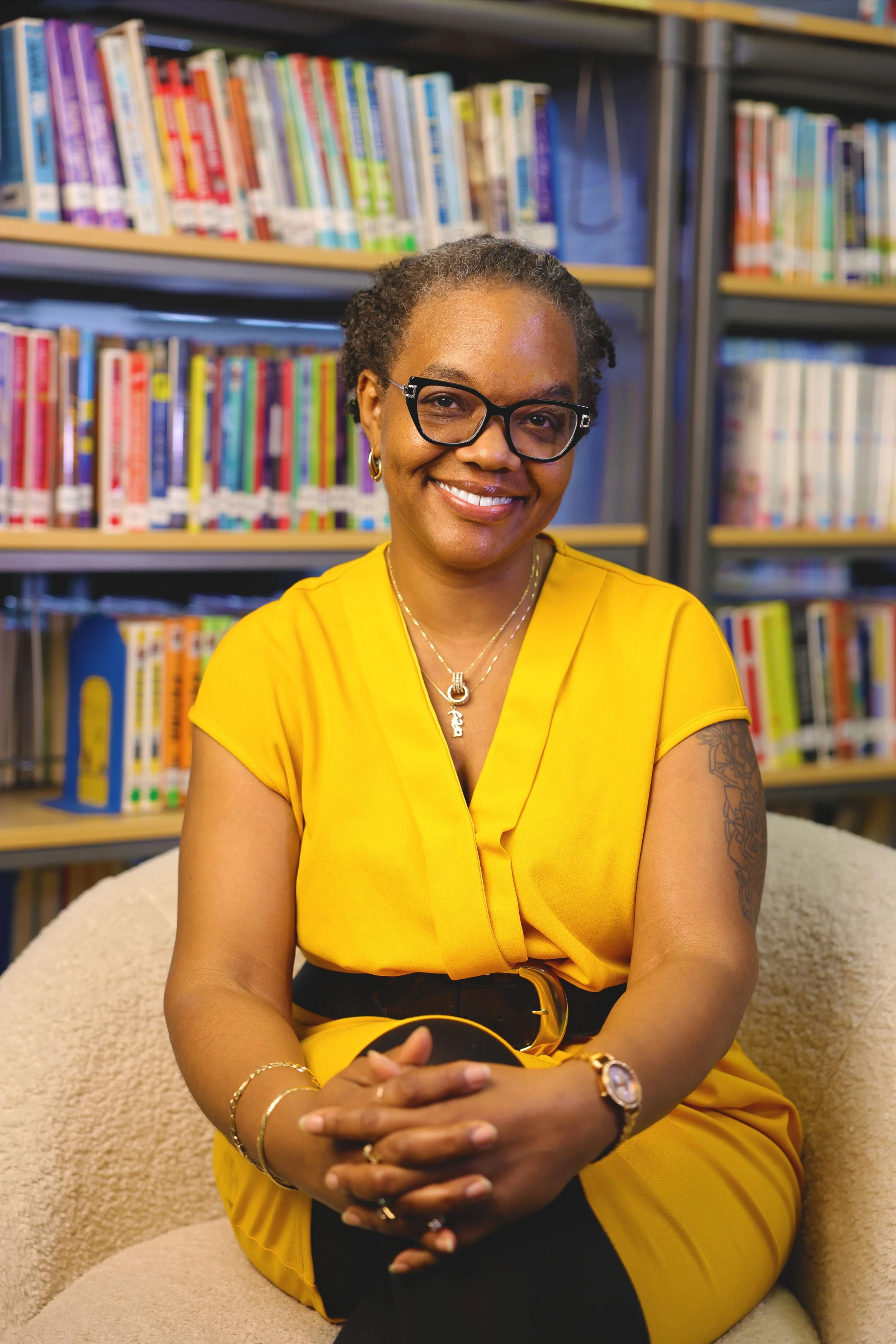 Elizabeth J. Harris, MSW, founder of Cynthy's Kids, Inc., smiling warmly in a yellow top while seated in front of a colorful bookshelf, representing her passion for education, community, and legacy.