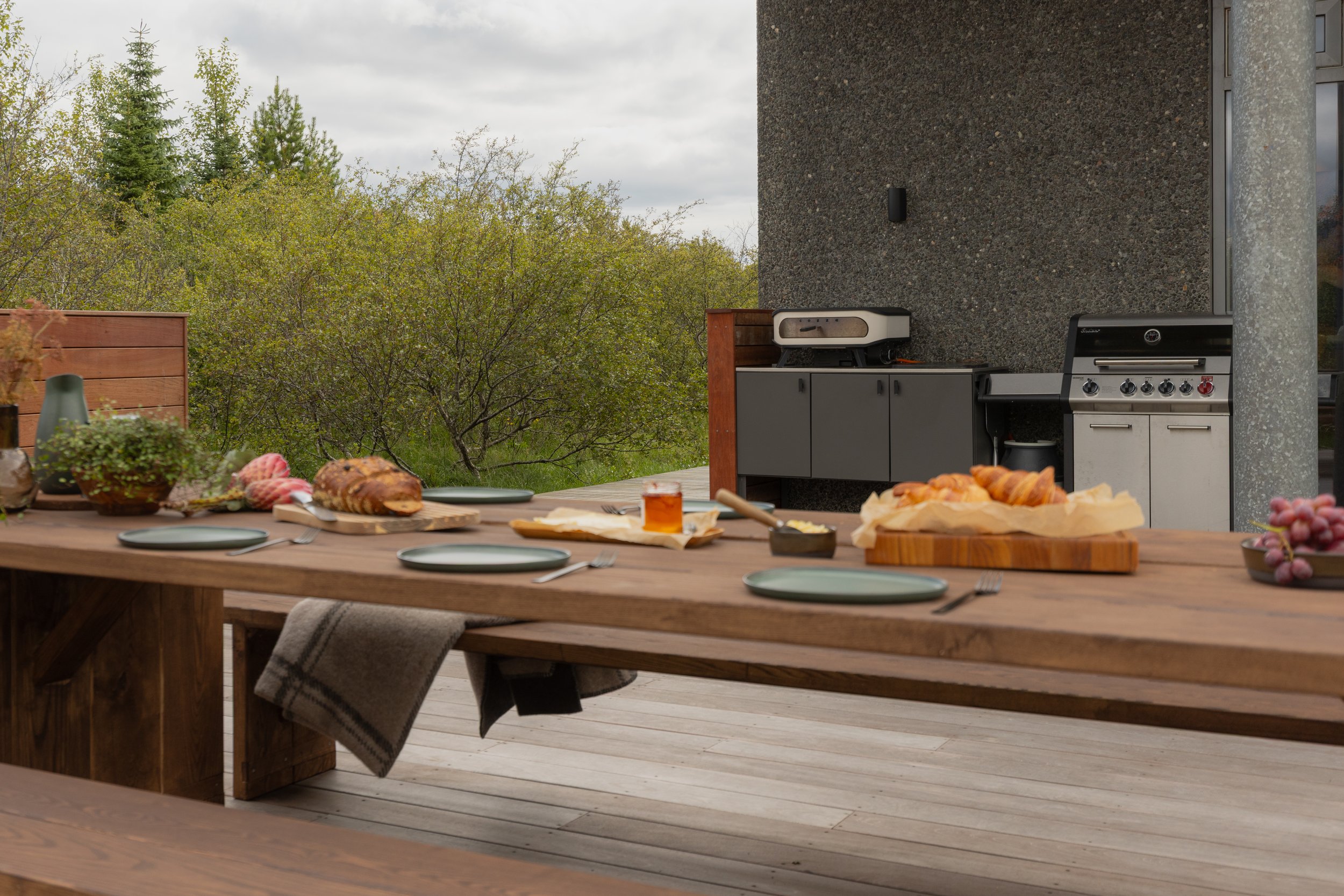 Outdoor patio with a large wooden dining table set with plates, silverware, bread, and grapes, alongside a charcoal grill, with trees and a cloudy sky in the background.