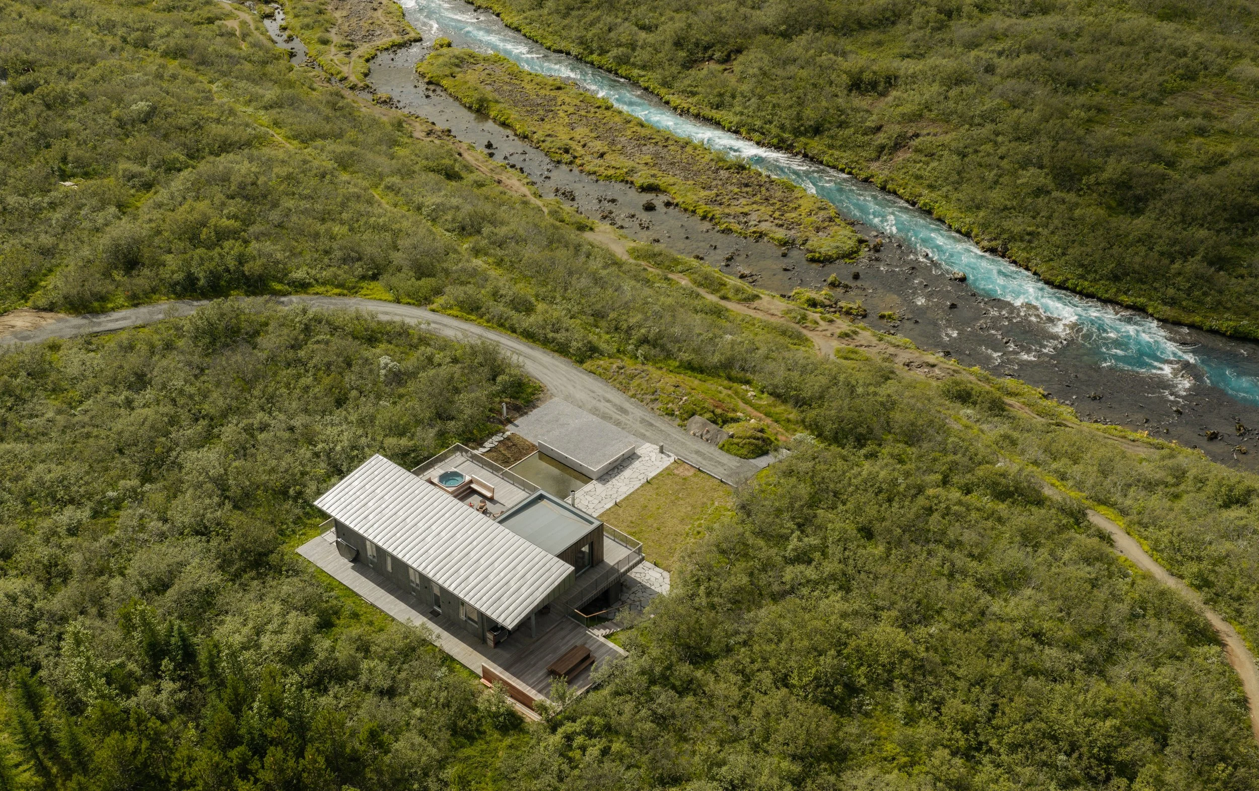 Aerial view of a modern house with a metal roof next to a winding river in a lush, green forest.