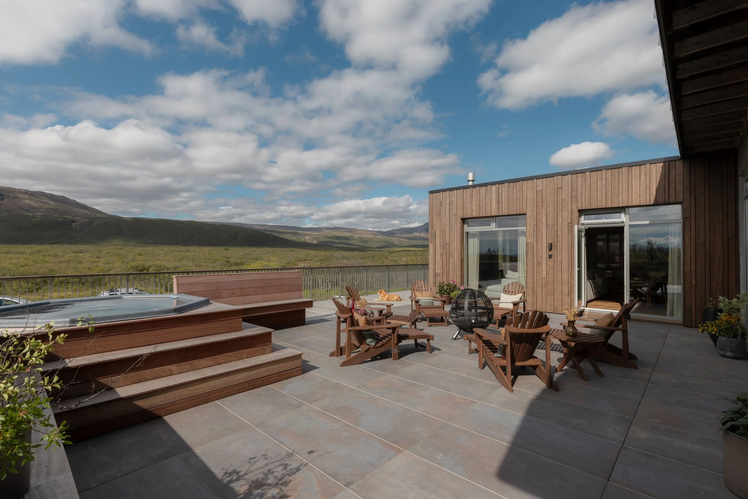A spacious outdoor patio overlooking rolling green hills and mountains under a partly cloudy sky. The patio features wooden furniture, a hot tub, potted plants, and large glass doors leading into a modern wooden house.