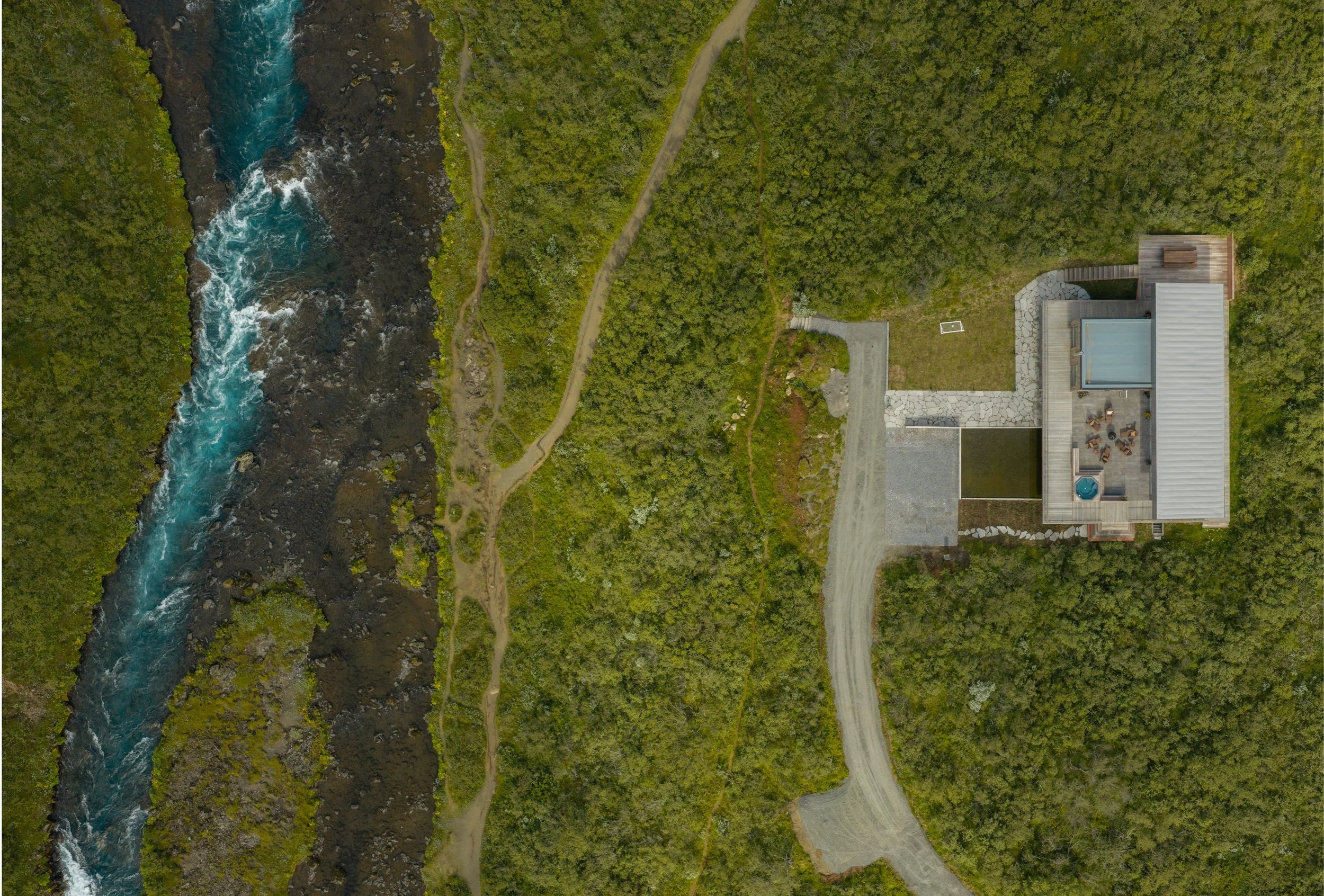 Aerial view of a landscape with a river on the left, trees, and a house with a patio on the right.