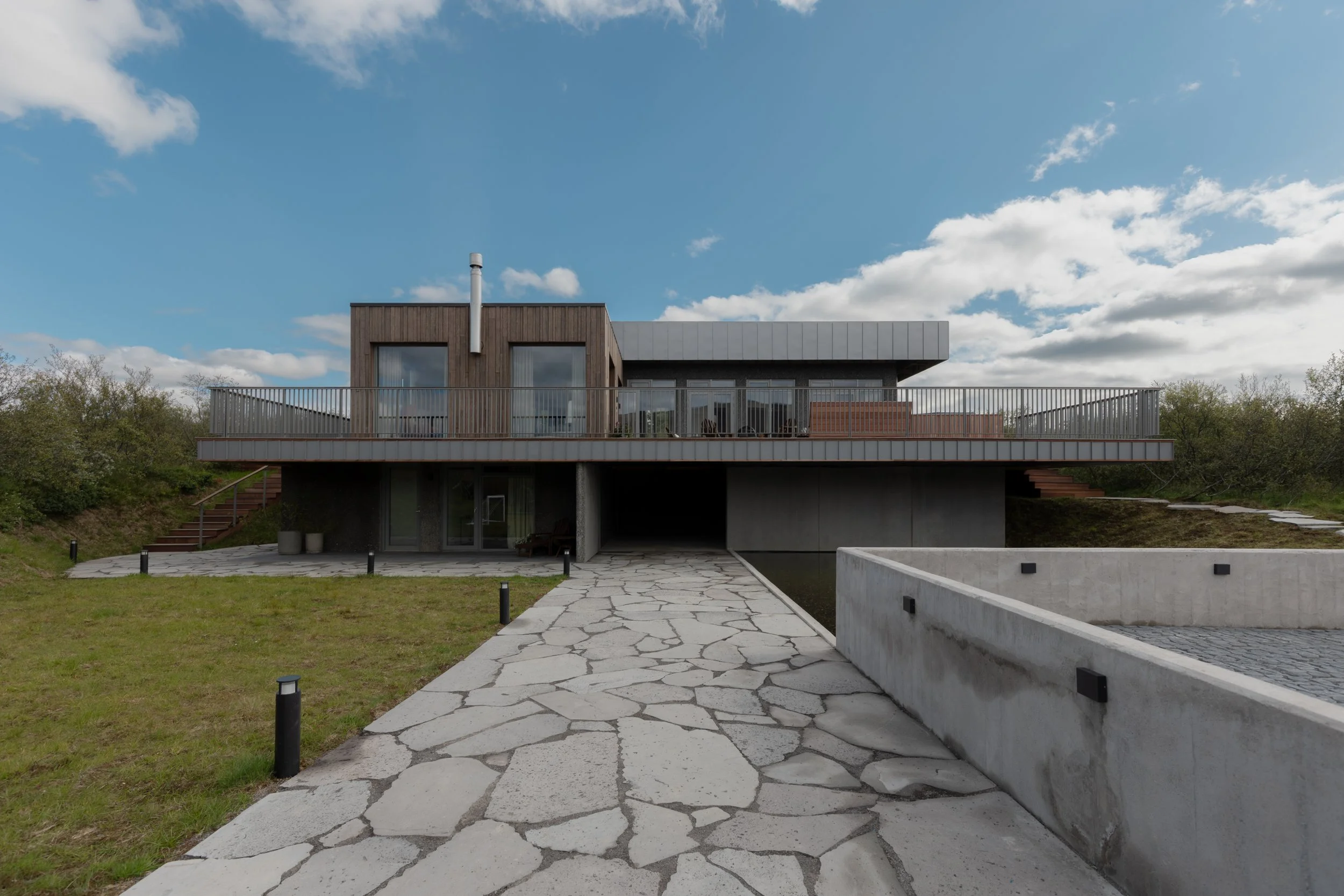 Modern two-story house with a flat roof, large windows, and a wooden exterior, surrounded by a lawn, trees, and a stone pathway leading to the entrance under a cloudy sky.