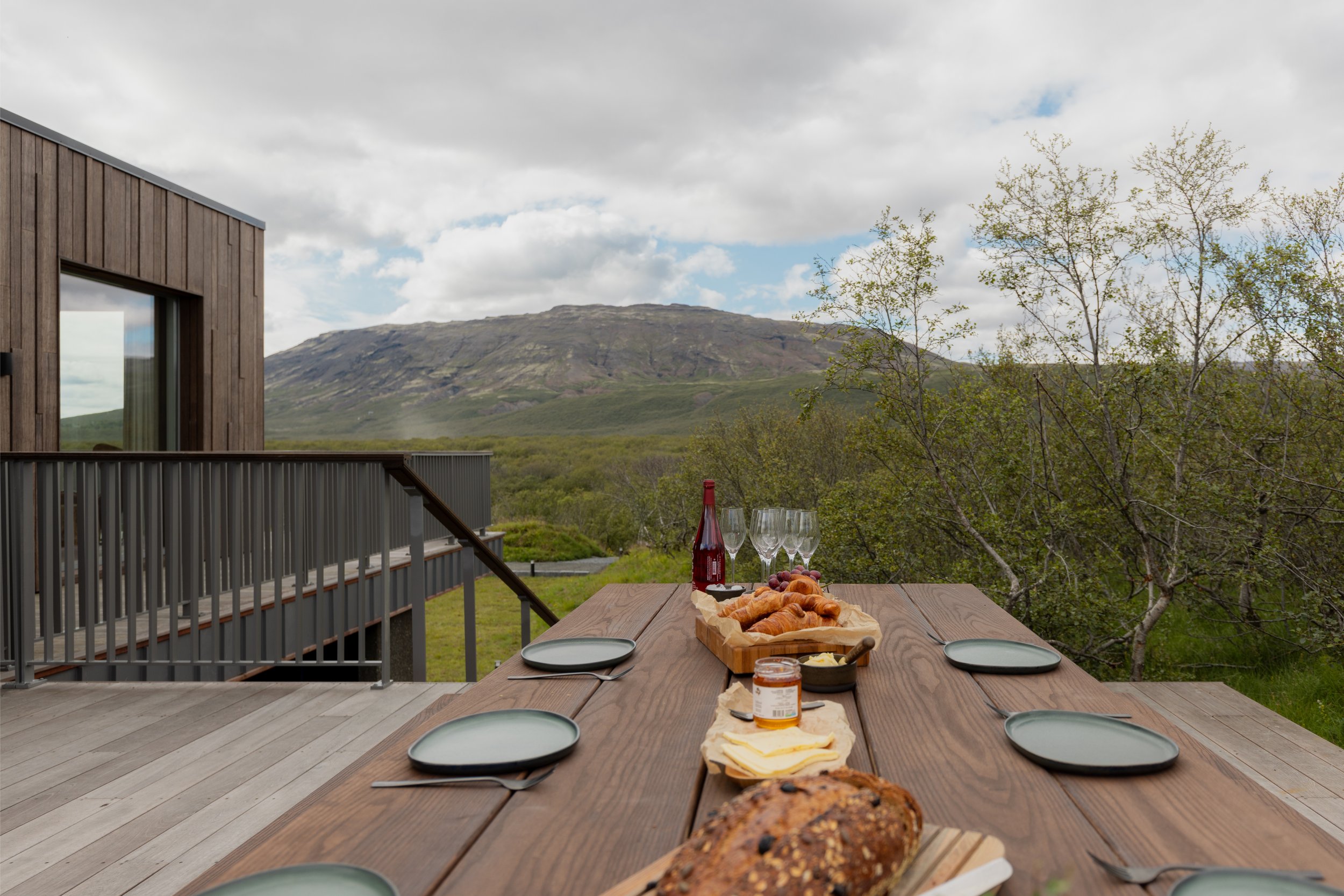An outdoor dining table set for a meal with a mountain view in the background. The table has bread, cheese, a jar of jam, a bottle of wine, wine glasses, and empty plates with utensils. There is a house behind the table with a wooden exterior and a r