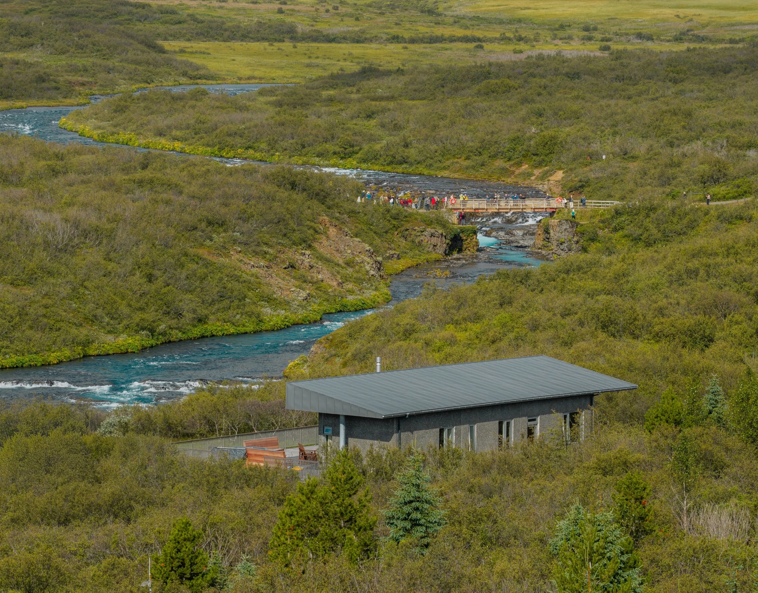 View of a river flowing through a lush green landscape with a bridge and several people walking on it. In the foreground, there is a building with a metal roof and a balcony.