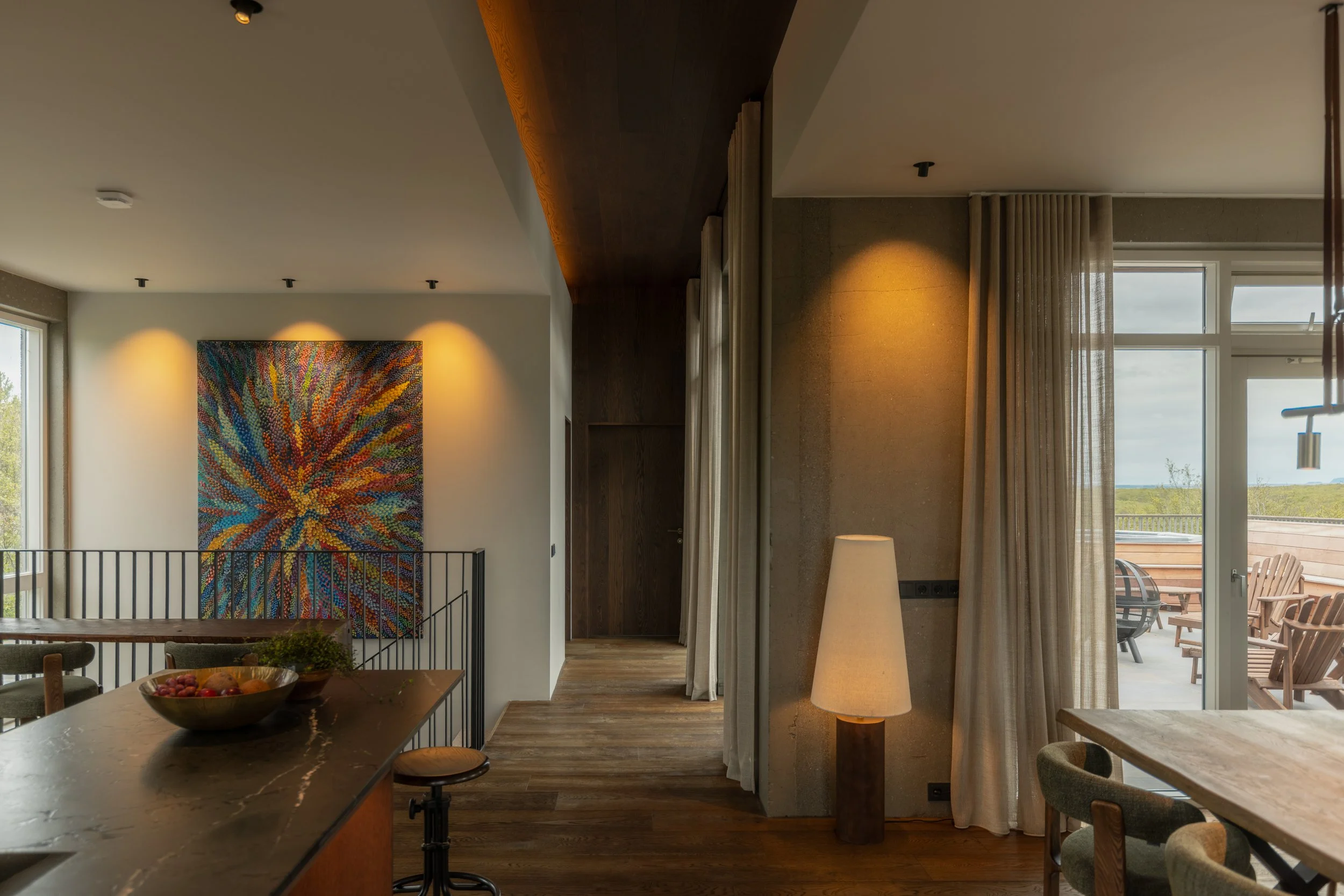 A modern interior living space with a large window and sliding glass door leading to a deck with outdoor chairs. The room features a colorful abstract wall art, a tall floor lamp, and curtains. The ceiling has recessed lighting, and a mixture of wood