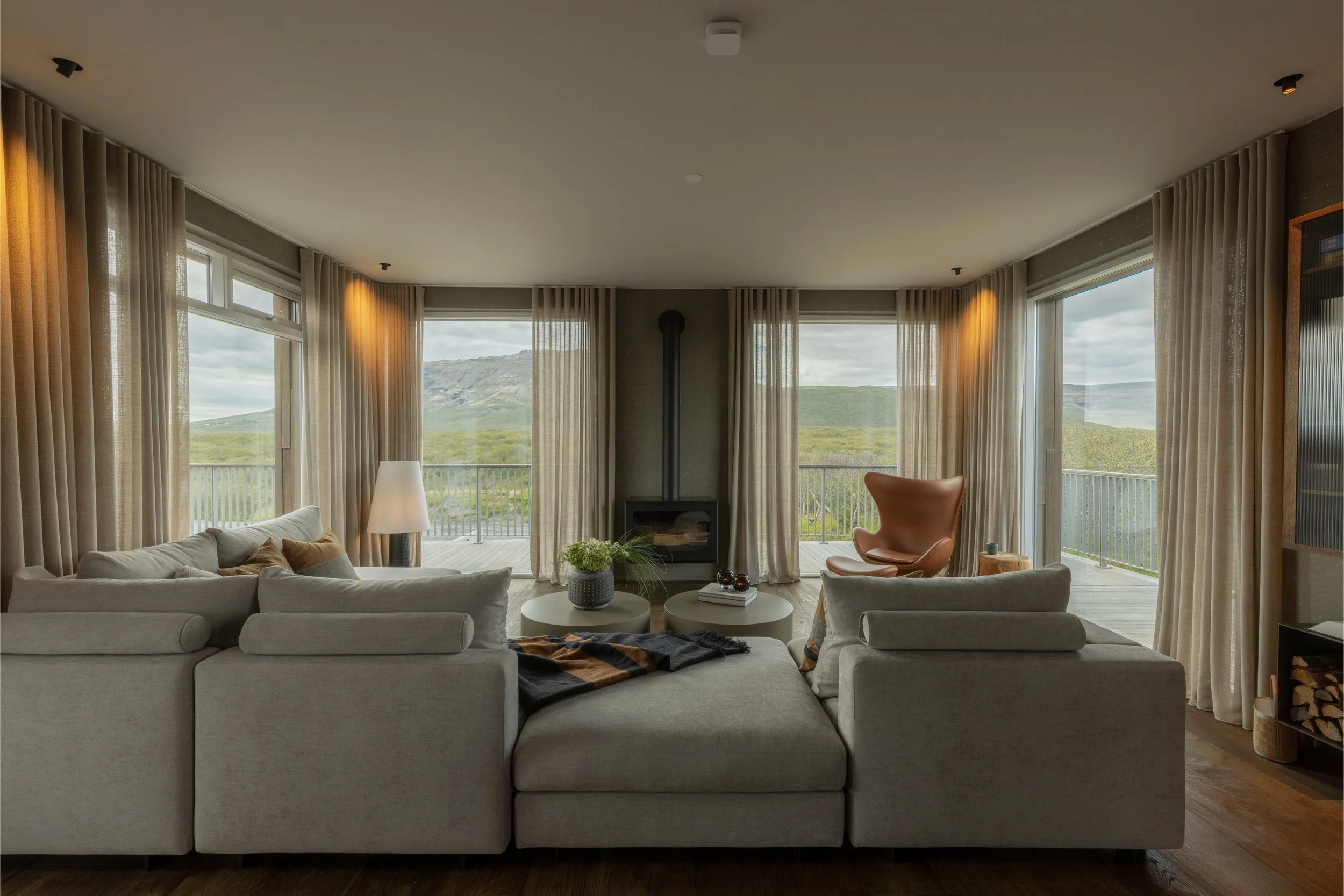 A living room with large windows showing a mountain landscape, beige curtains, white sofa, orange armchair, coffee tables, and a fireplace.