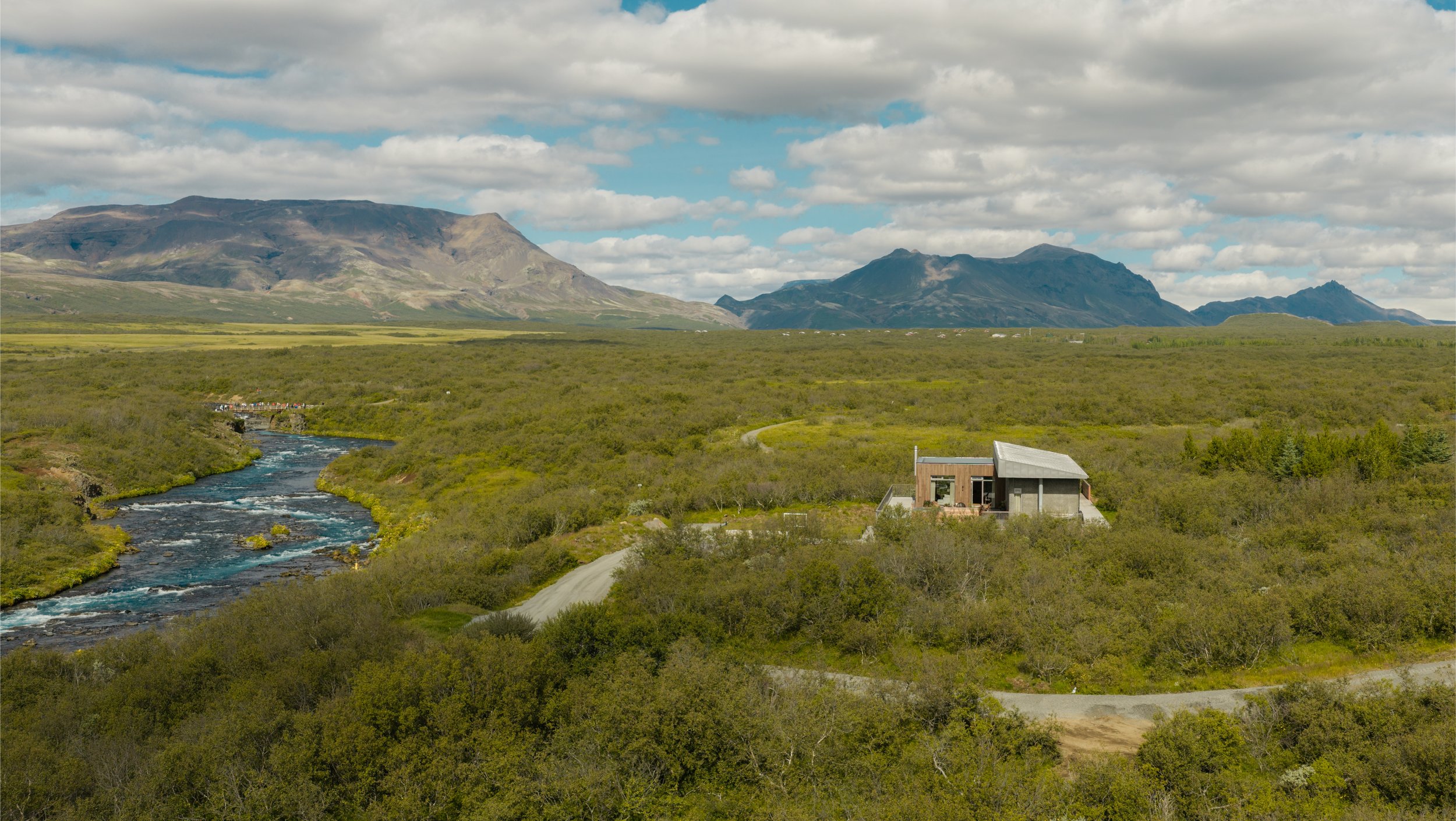 A scenic landscape with a river flowing through green vegetation, a modern house, and mountains in the background under a partly cloudy sky.