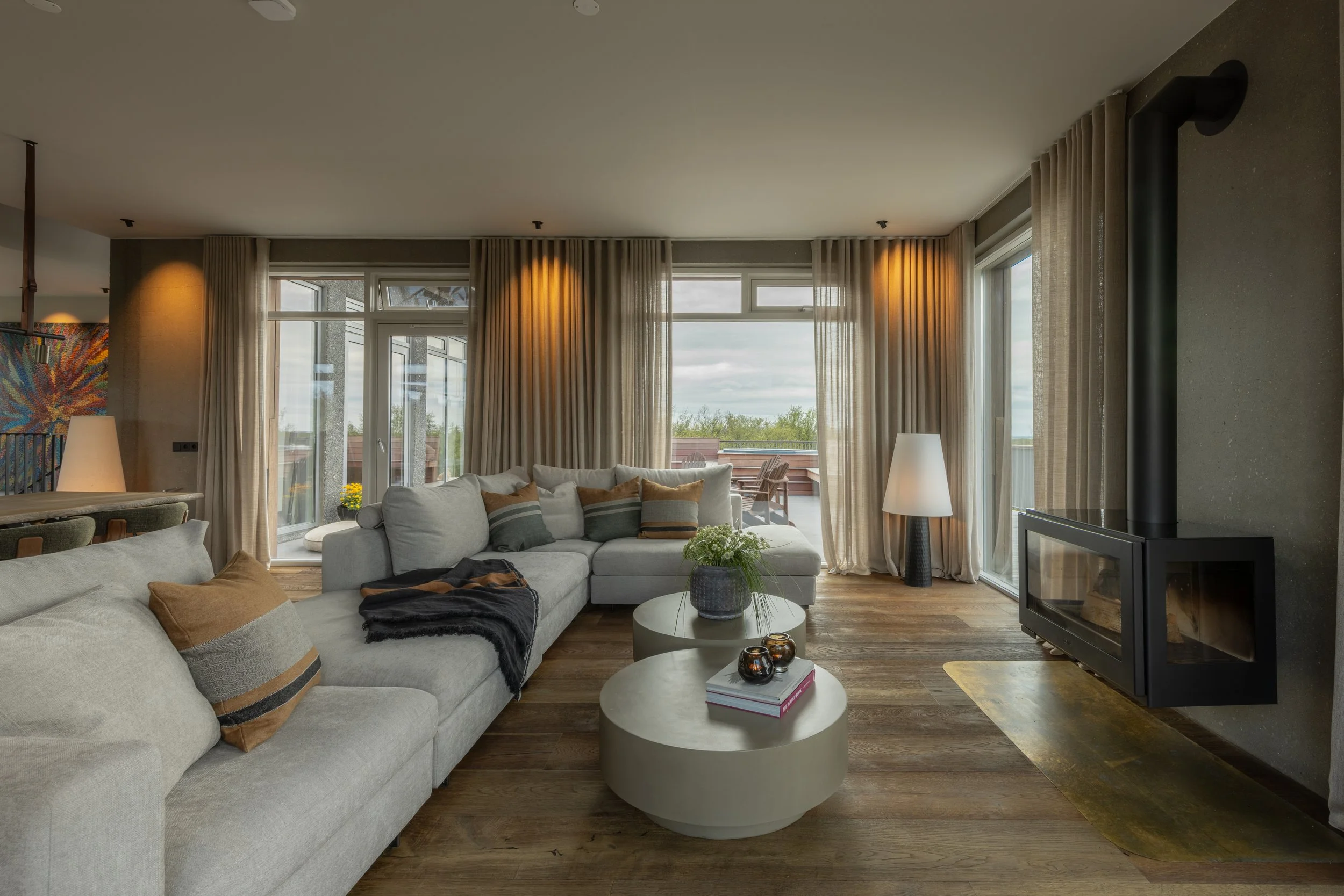 Living room with light-colored sofa, throw pillows, coffee tables, a fireplace, and large windows with beige curtains, overlooking an outdoor balcony.