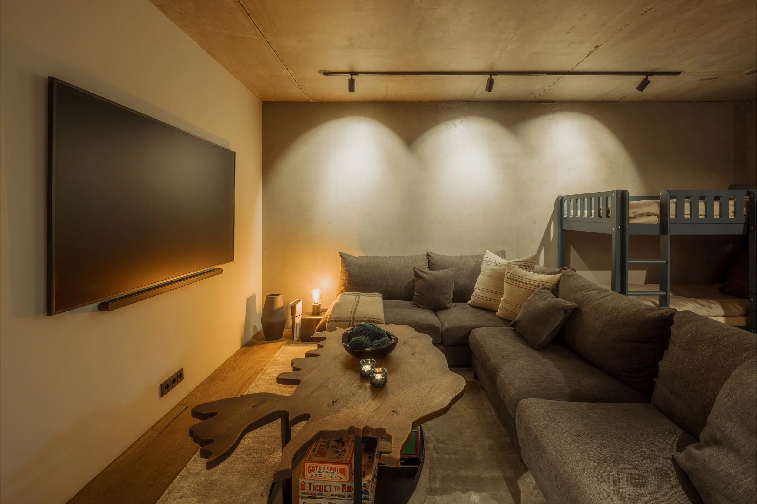 A cozy living room with a large gray sectional sofa with beige and striped pillows. A wooden coffee table with a sculpted edge holds candles and a decorative bowl. There's a large flat-screen TV mounted on a cream-colored wall. A small side table wit