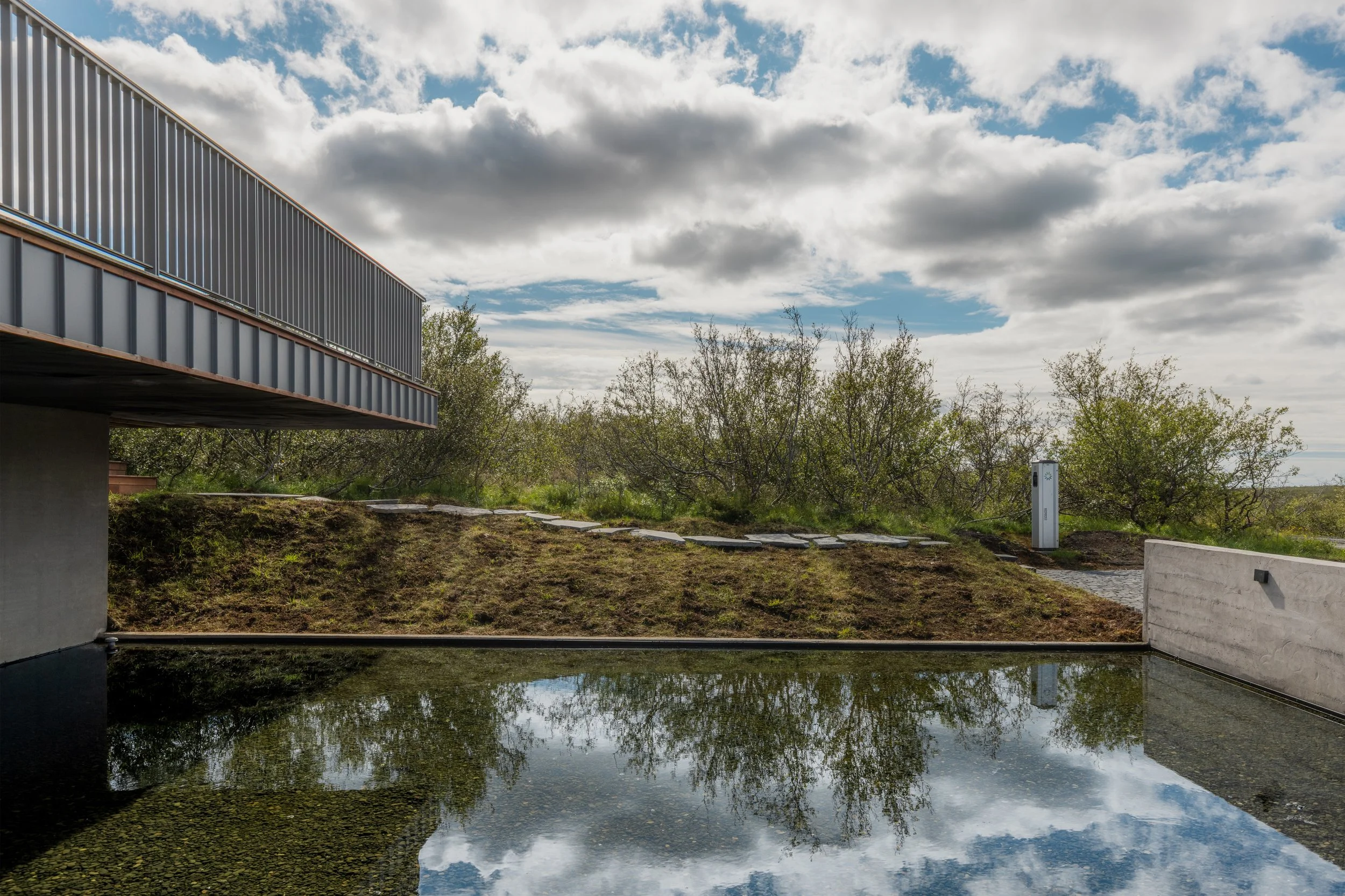 A modern building with a metal railing balcony overlooks a pond, with trees in the background and a partly cloudy sky overhead.