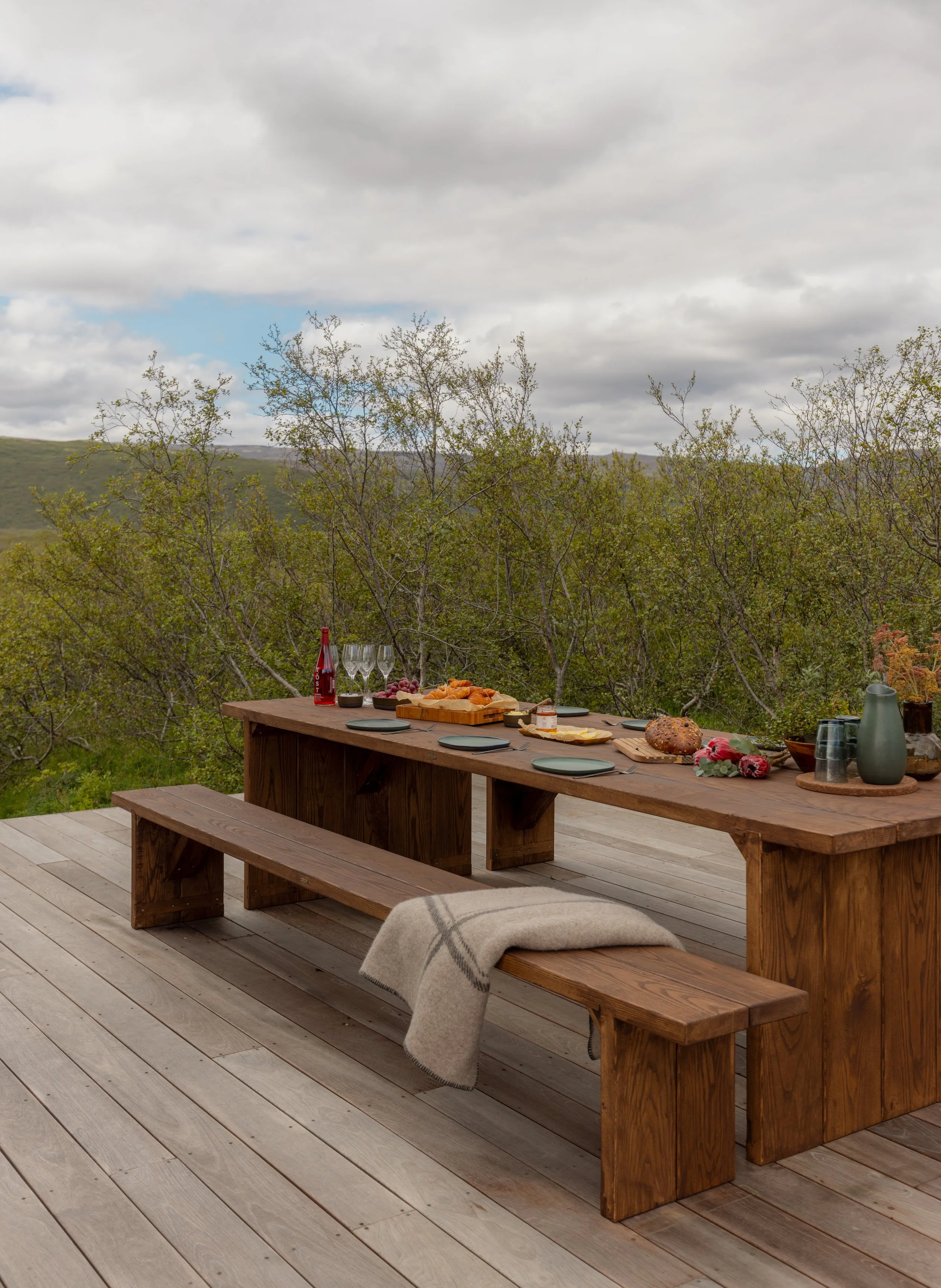 Outdoor wooden dining table with a bench and stool, set with wine glasses, plates, a bottle, and food, on a wooden deck with a green hilly landscape and cloudy sky in the background.