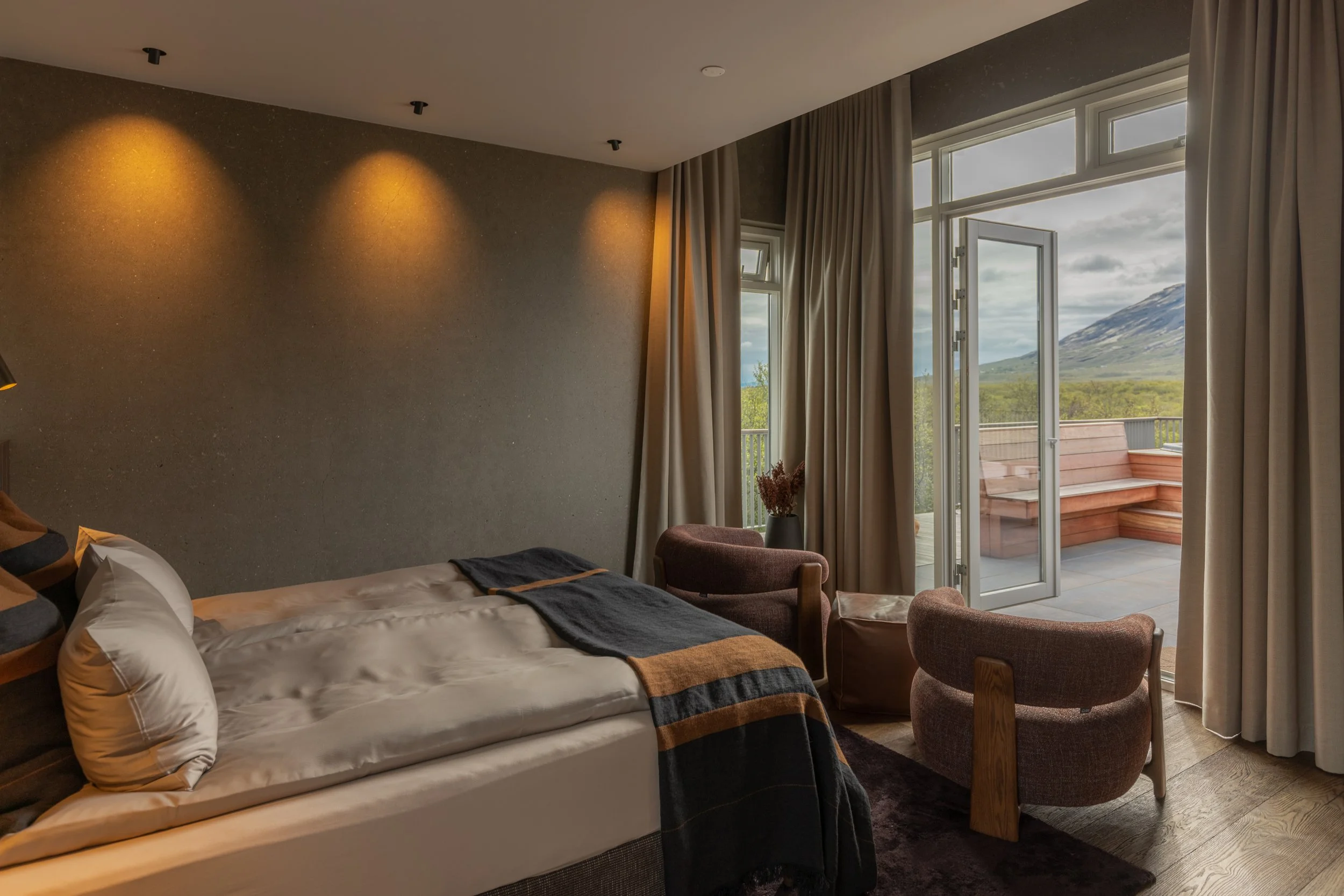 A bedroom with a bed, throw pillows, table lamp, curtain-draped large window opening to a balcony with mountain view.