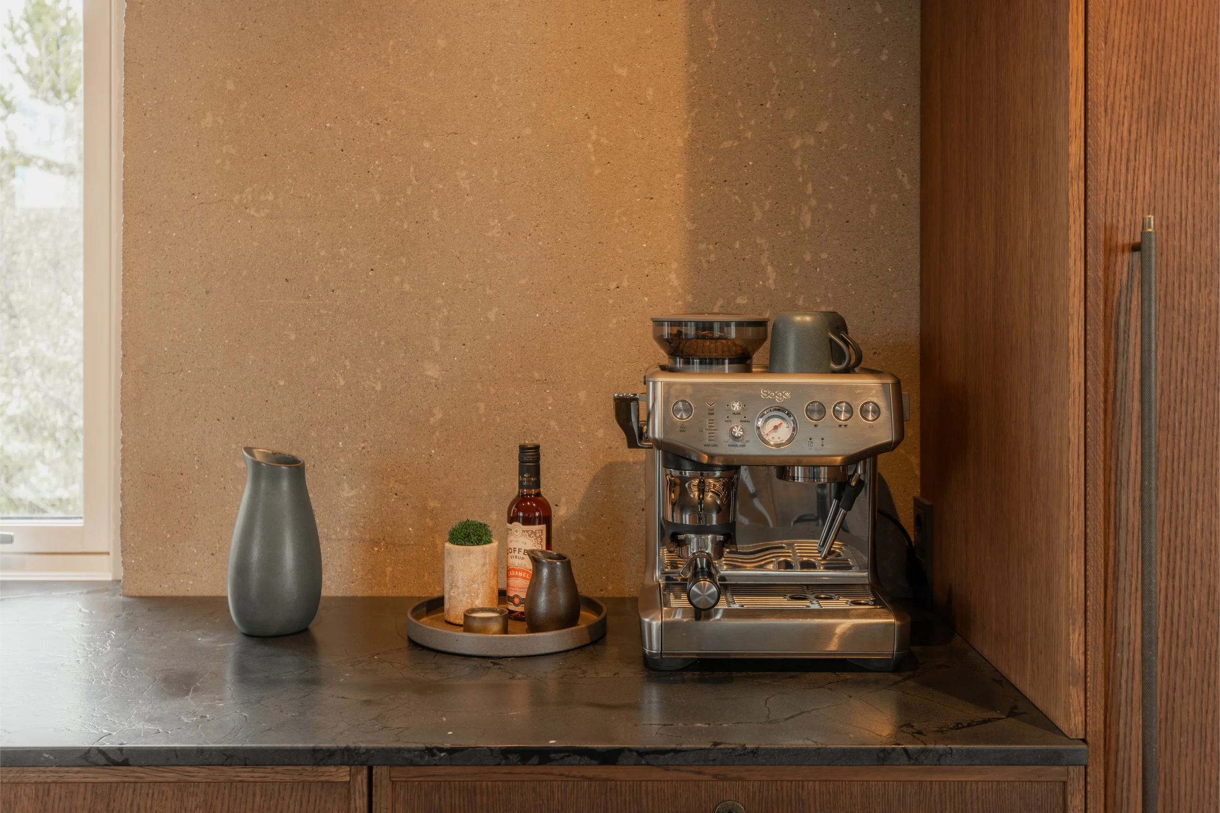 A kitchen countertop with a gray vase, a bottle, and decorative items, positioned in front of a window, with a coffee machine against a textured beige wall and a wooden cabinet to the side.