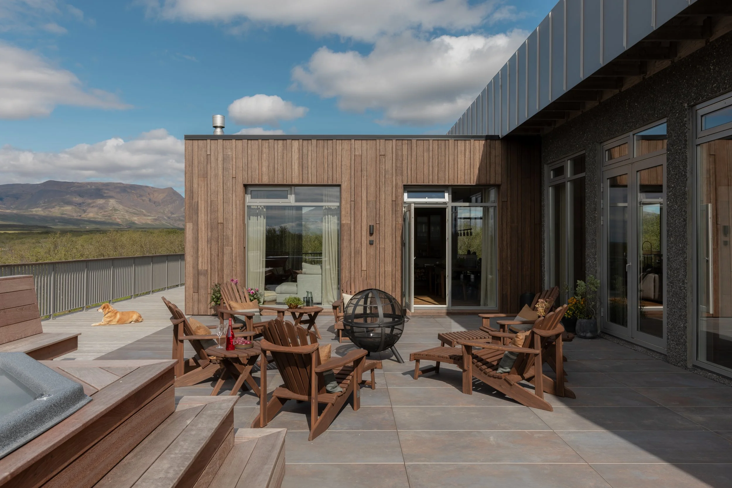 An outdoor patio area with wooden chairs, a fire pit, and a dog lying on the ground. In the background, there is a house with wooden and glass exterior walls and large windows, set against a backdrop of mountains and a blue sky with clouds.