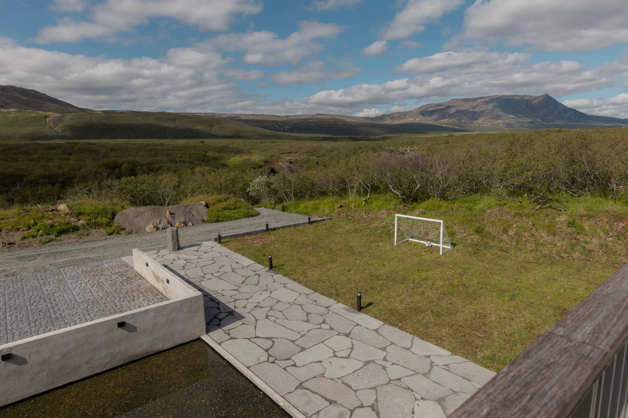 Landscape view of green hills, mountains in the distance, scattered clouds in the sky, and a small soccer goal on a grassy area with a paved walkway.