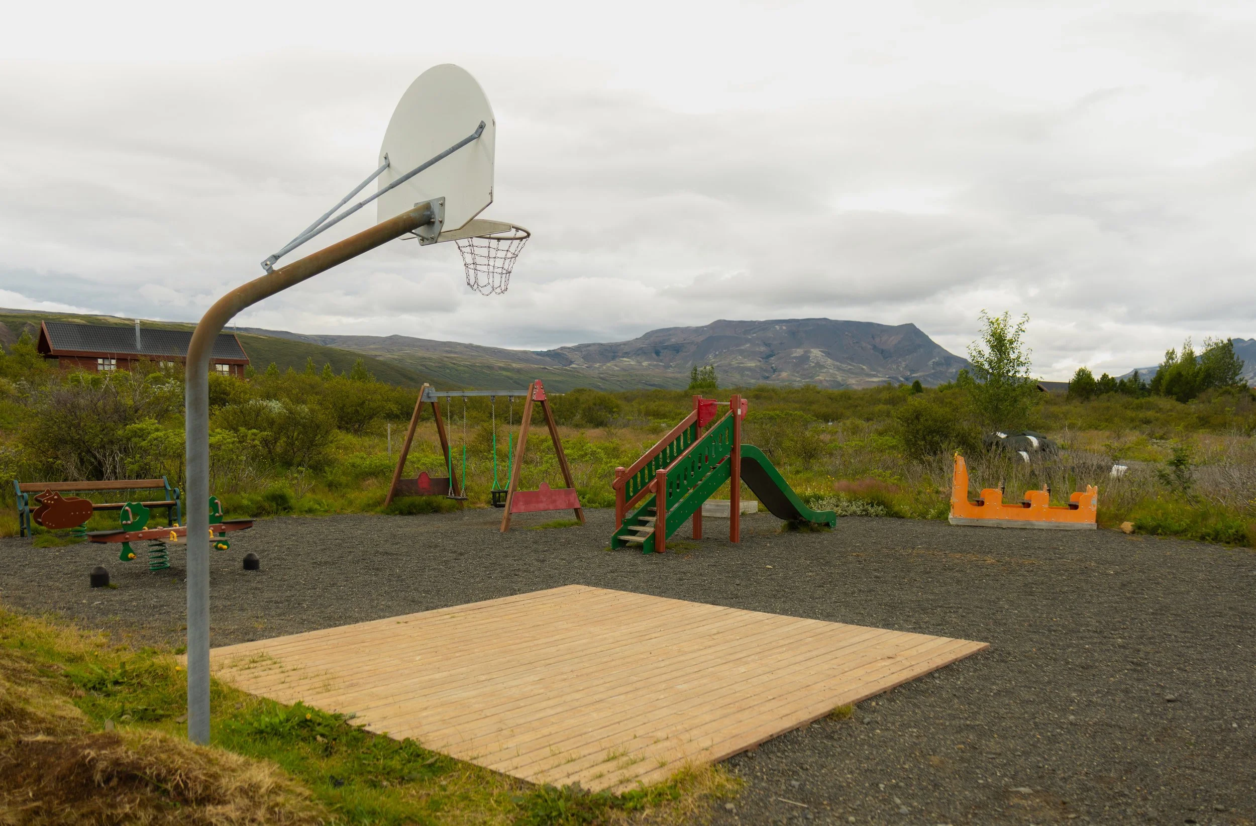 An empty playground with a basketball hoop, swings, slide, and rocking animals, set against a backdrop of mountains and cloudy sky.