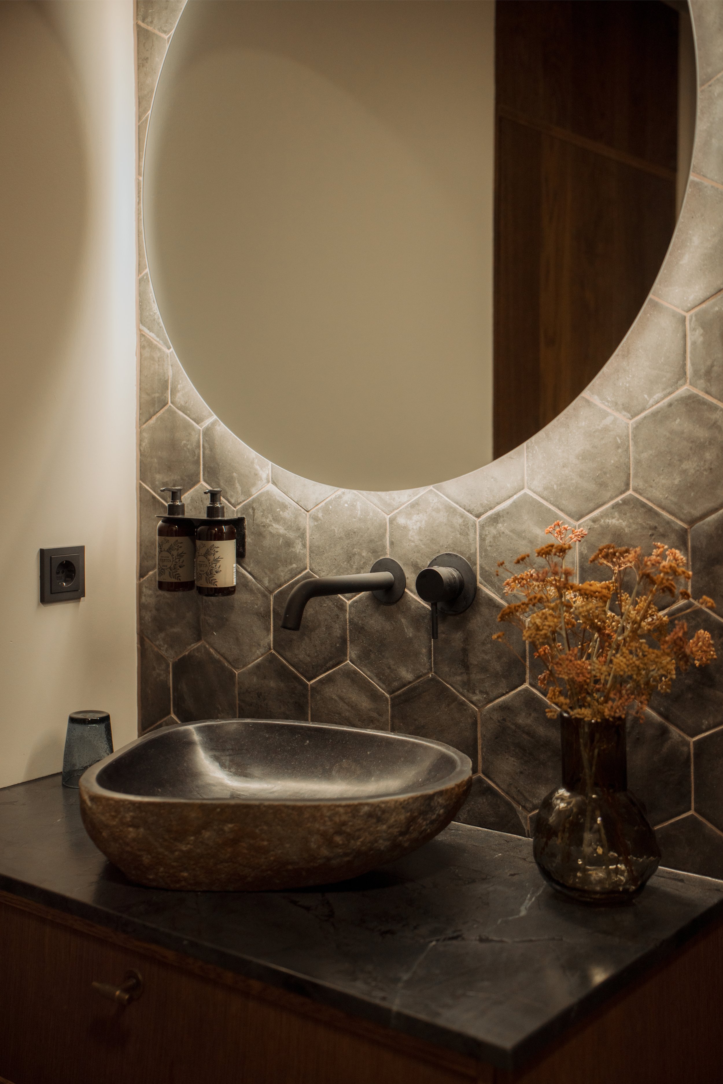 A bathroom sink vanity with a dark stone countertop, a textured ceramic vessel sink, a bronze faucet, a hexagon tile wall in shades of gray, a round mirror, wall-mounted soap dispensers, and a vase with dried flowers.