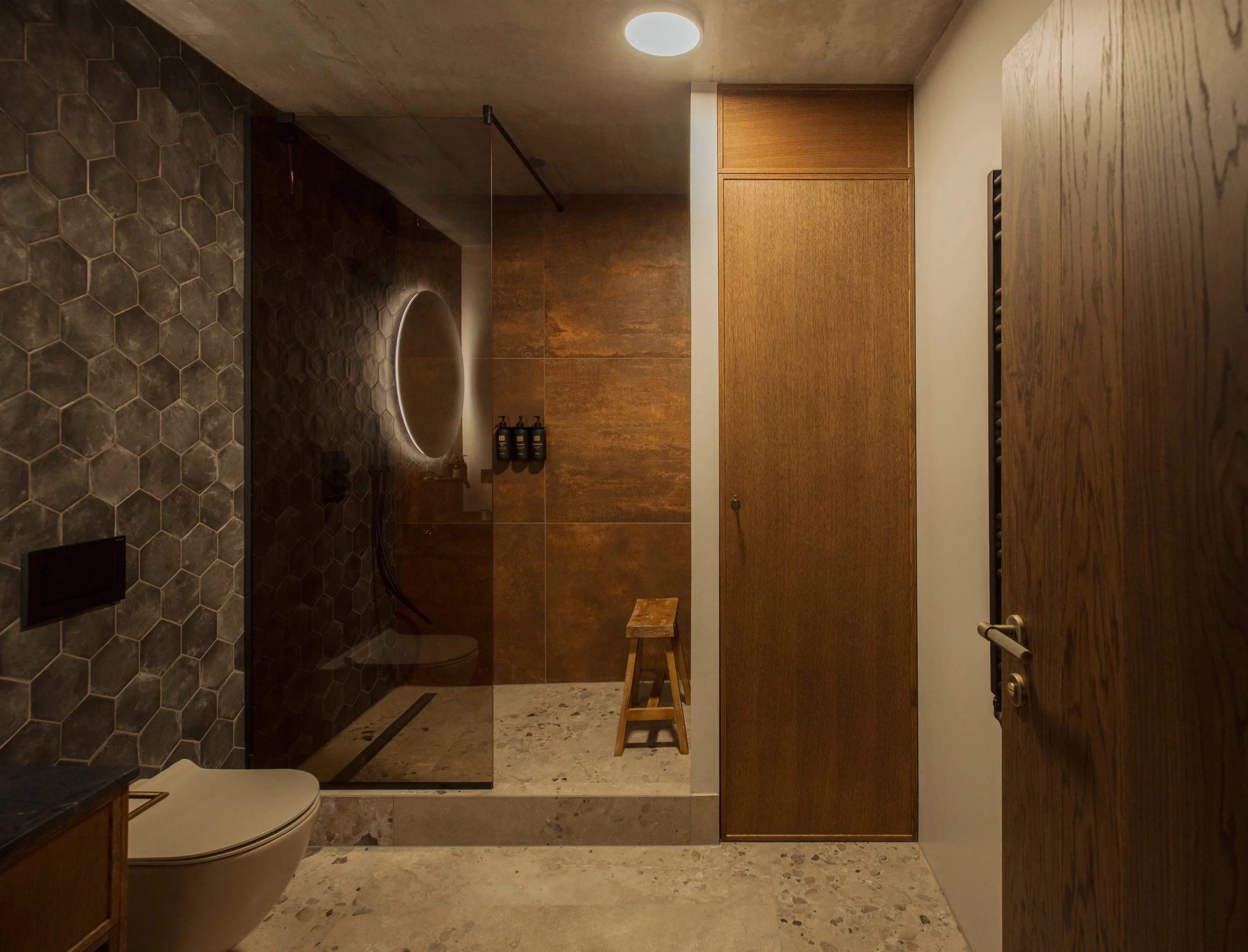 Modern bathroom with concrete ceiling, hexagonal tile wall on the left, wooden cabinets on the right, a walk-in shower with a glass door, a small stool inside the shower, and a white toilet in the foreground.
