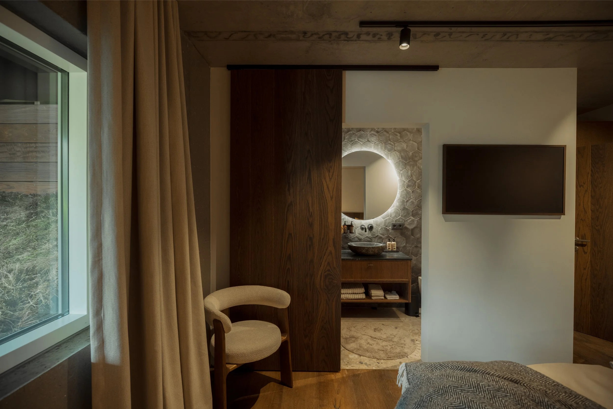 Interior of a modern room with a window and beige curtains on the left, a beige upholstered chair, a large flat-screen TV mounted on the wall, a mirror with circular lighting, a stone sink, and wooden furniture with shelves holding towels.