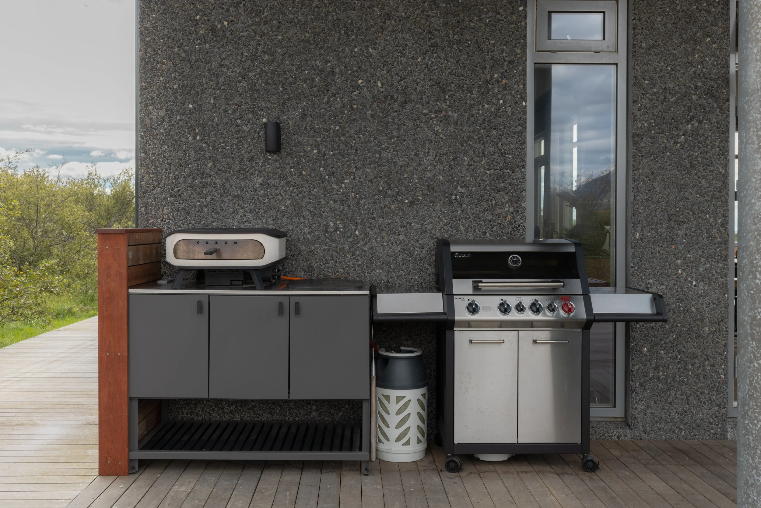 Outdoor kitchen with a gas grill and a small oven on a deck with a stone wall background.