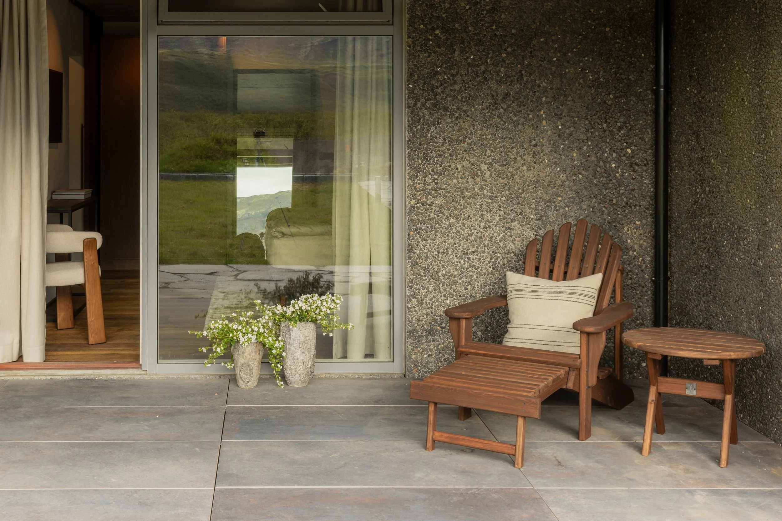 A patio with two wooden chairs near a wall, a sliding glass door, and outdoor plants.