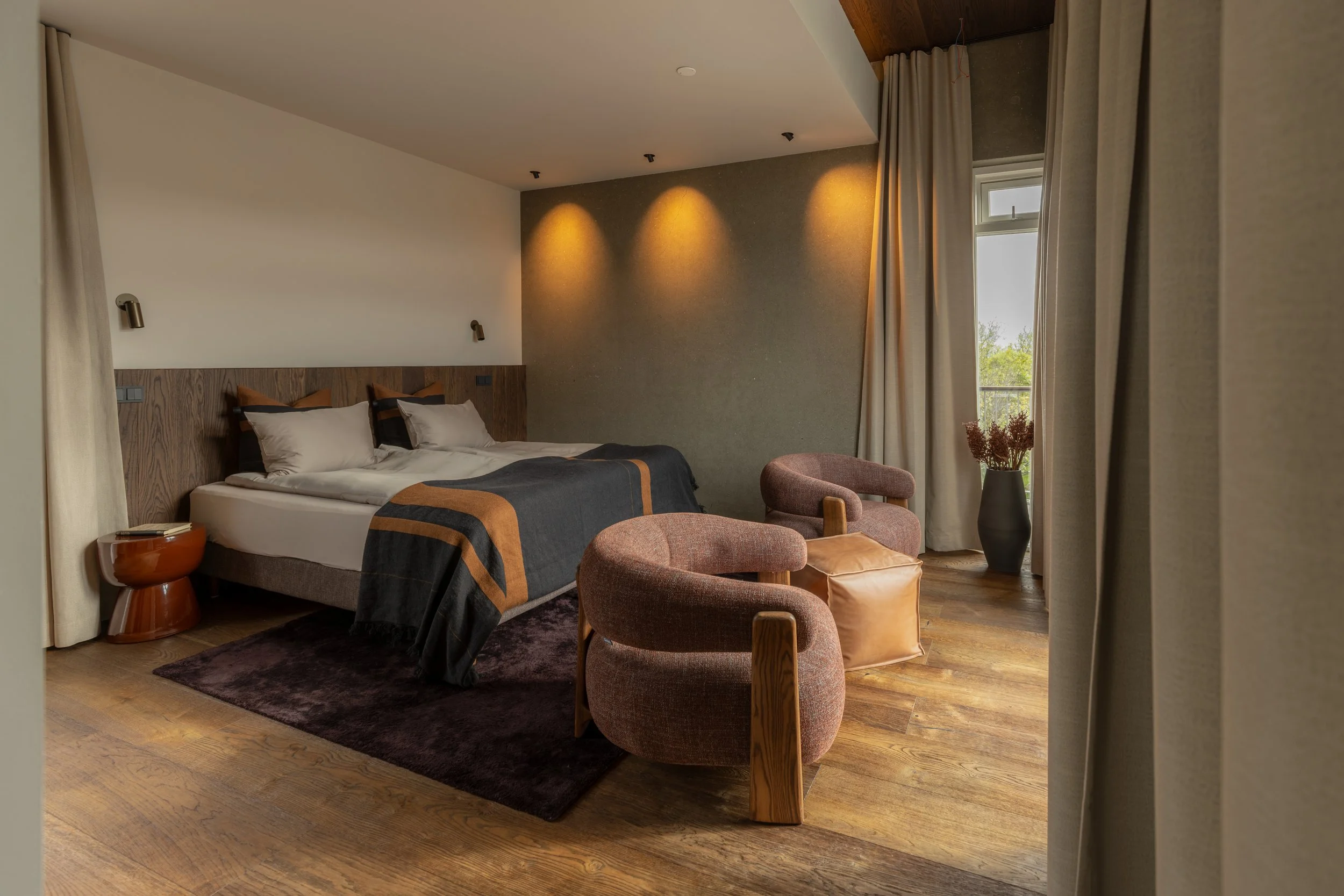 Interior of a modern bedroom with a bed, two chairs, a side table, and large window with beige curtains, wood flooring, and warm lighting.