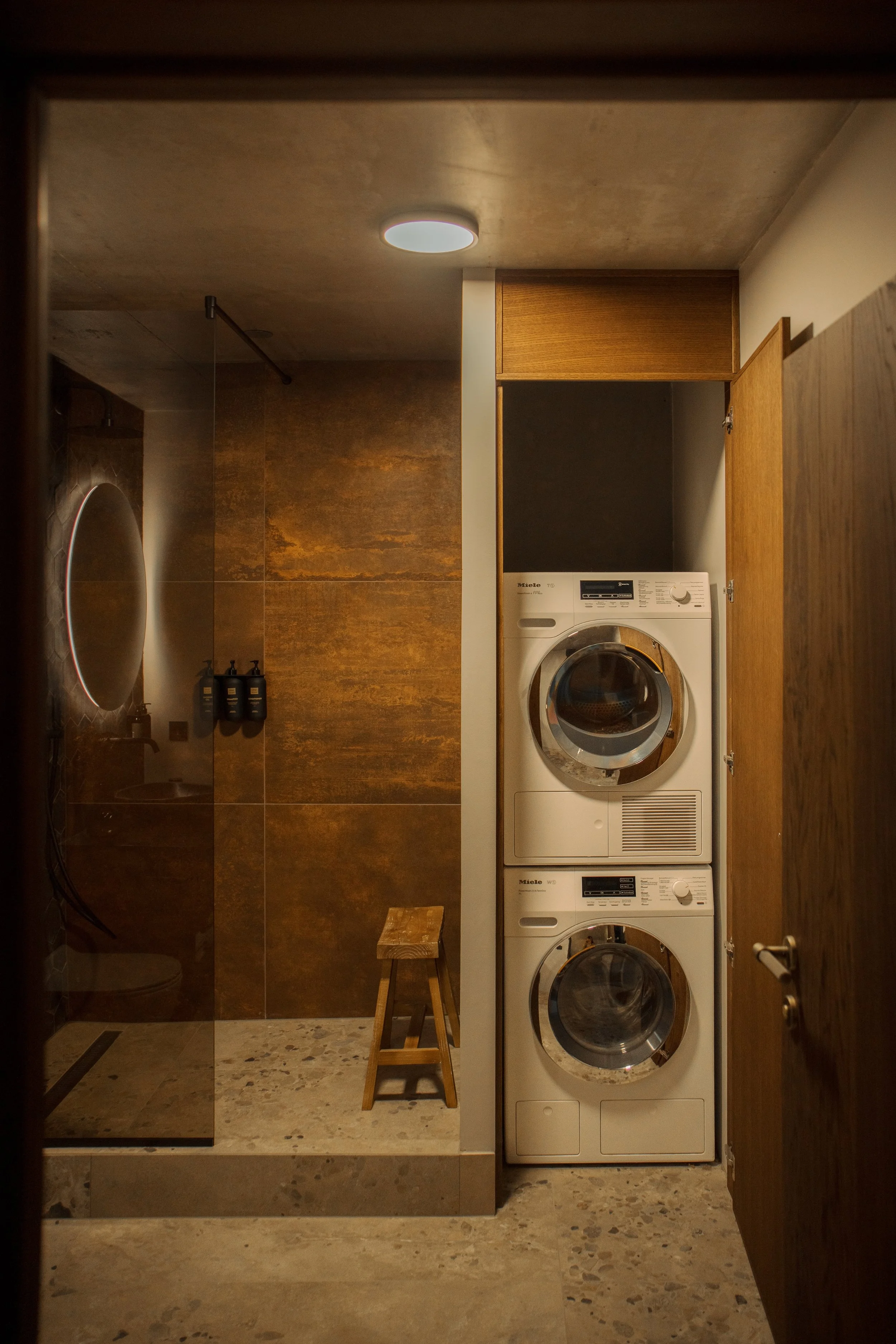 A small laundry room with a stacked washer and dryer next to a wooden cabinet, a wooden stool, and a round mirror on the wall. The room has earth-toned tiles and a wooden door.