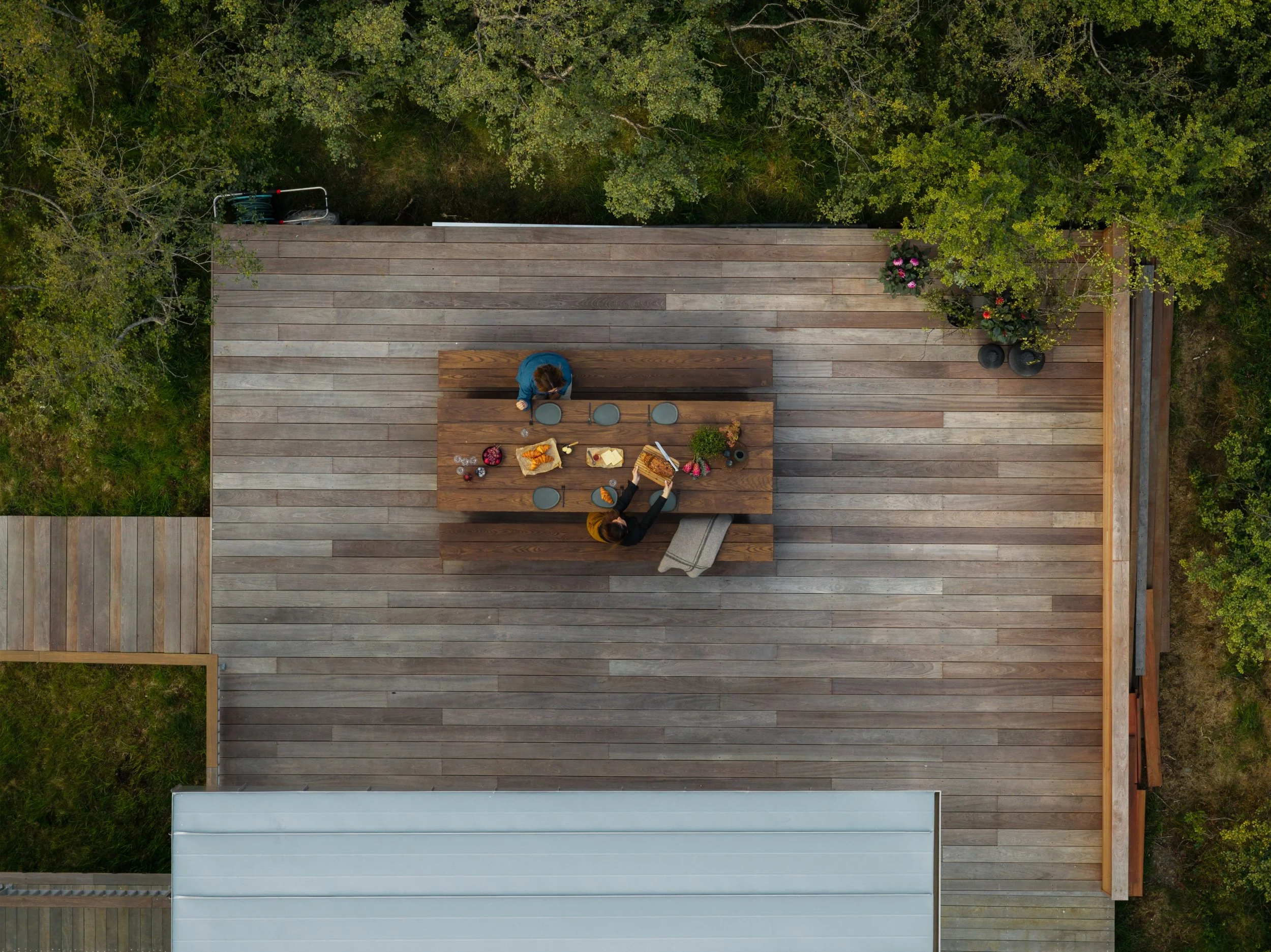 Bird's eye view of an outdoor wooden deck with two people sitting at a table, enjoying food and drinks, surrounded by greenery and potted plants.