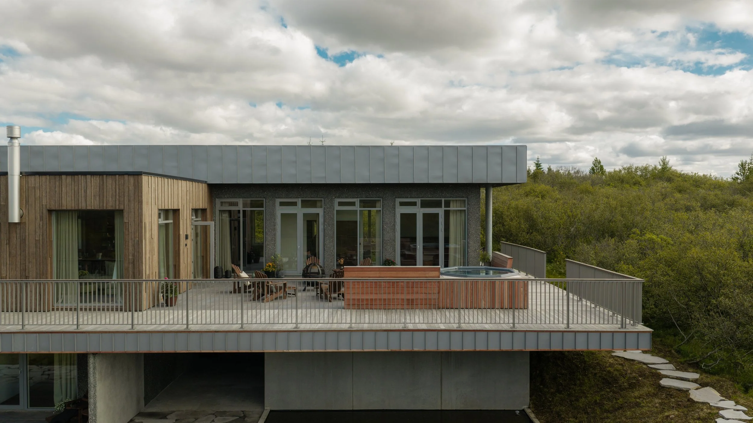 Modern multi-story house with wooden and gray exterior, large glass windows, balcony with outdoor furniture, hot tub, and surrounding greenery under cloudy sky.
