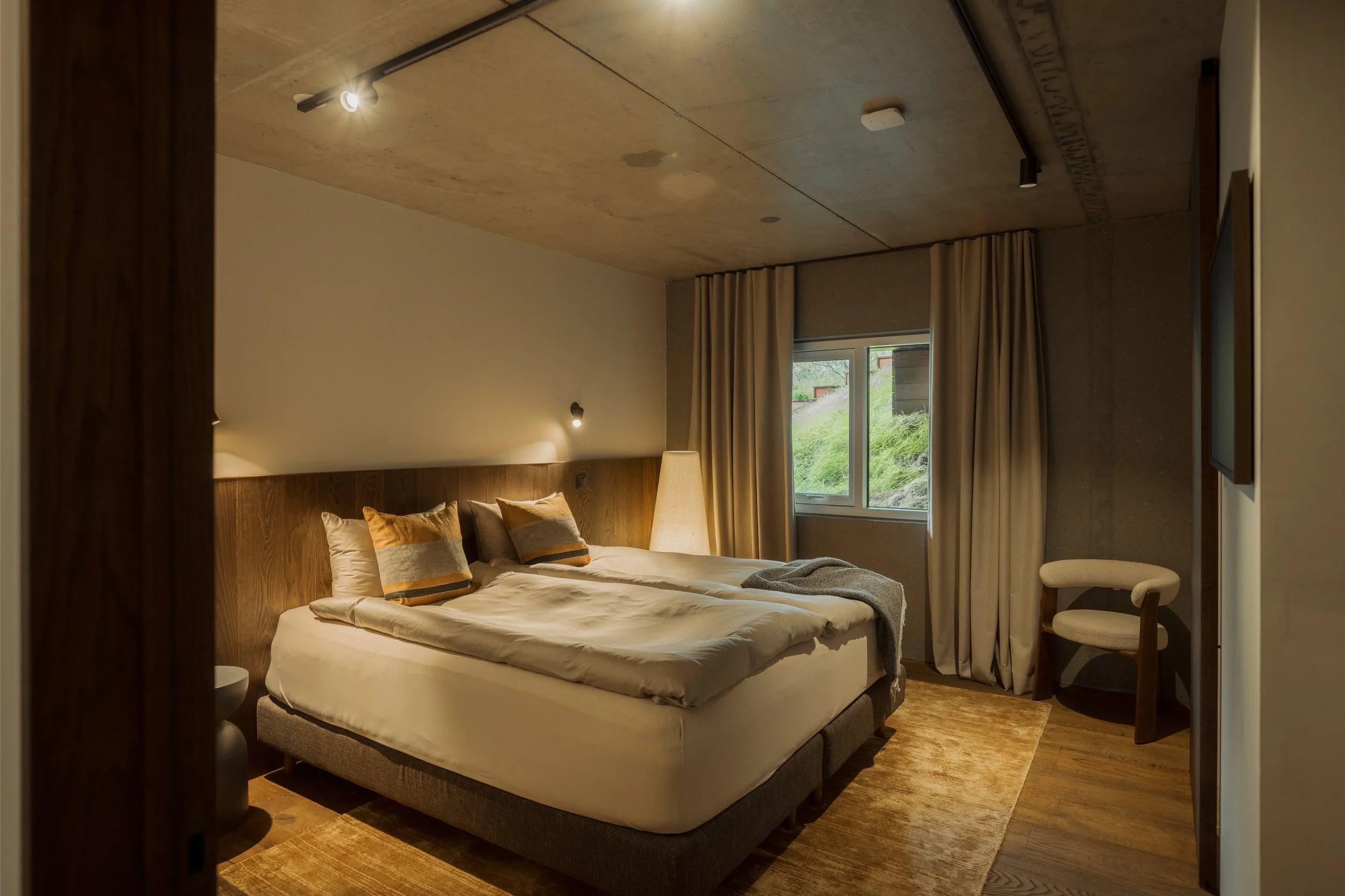 A cozy bedroom with a large bed, beige and brown pillows, a white blanket, and a wooden headboard. There's a tall floor lamp beside the bed, a window with beige curtains, and a chair in the corner. The ceiling is concrete with track lighting, and the room has wooden floors.
