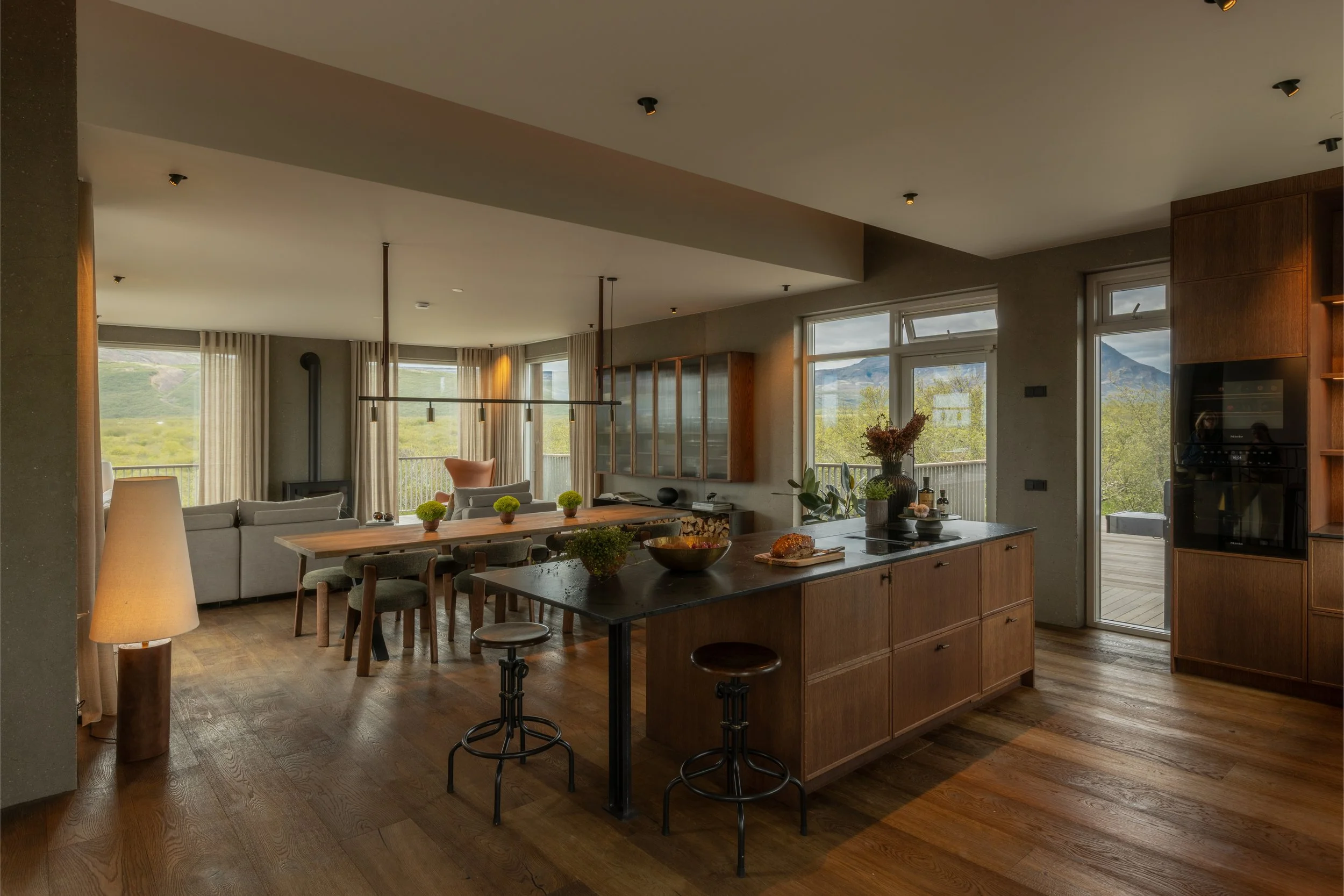 Open-concept living space and kitchen with large windows showing a mountainous landscape, wooden furniture, a dining table with chairs, a kitchen island with stools, and decorative plants.